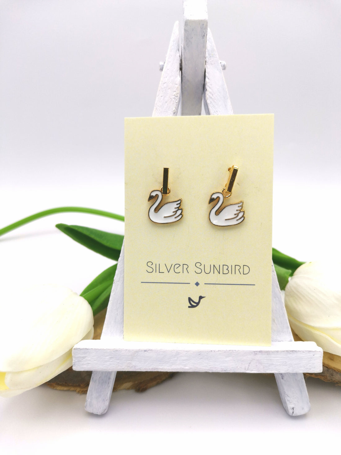 Graceful Swan Earrings by Silver Sunbird are displayed on a small easel, featuring gold-plated enamel swan charms. The minimal background includes white and green elements, with "Silver Sunbird" printed on the card to emphasize this stunning handmade jewelry.