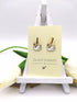 Graceful Swan Earrings by Silver Sunbird are displayed on a small easel, featuring gold-plated enamel swan charms. The minimal background includes white and green elements, with "Silver Sunbird" printed on the card to emphasize this stunning handmade jewelry.