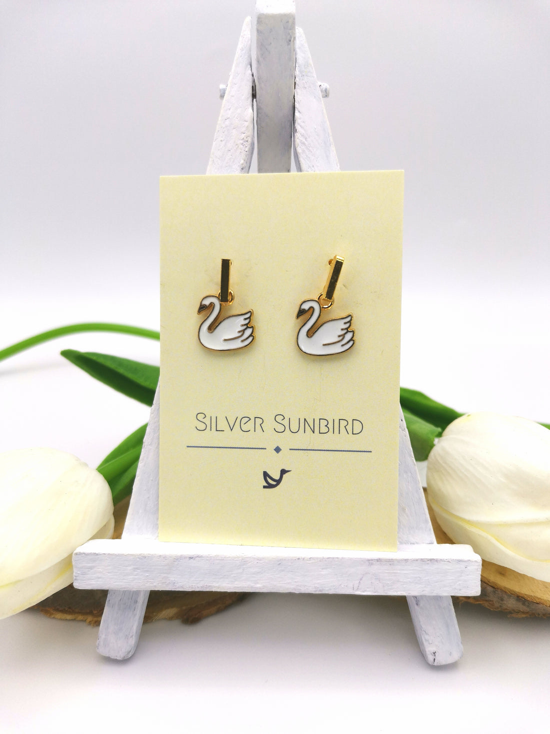 A pair of Graceful Swan Earrings with gold plated enamel swan charms, by Silver Sunbird, is elegantly displayed on a small easel. The card displays the brand name and delicate bird logo, while white flowers and a green leaf create a graceful backdrop highlighting the handmade jewelry&