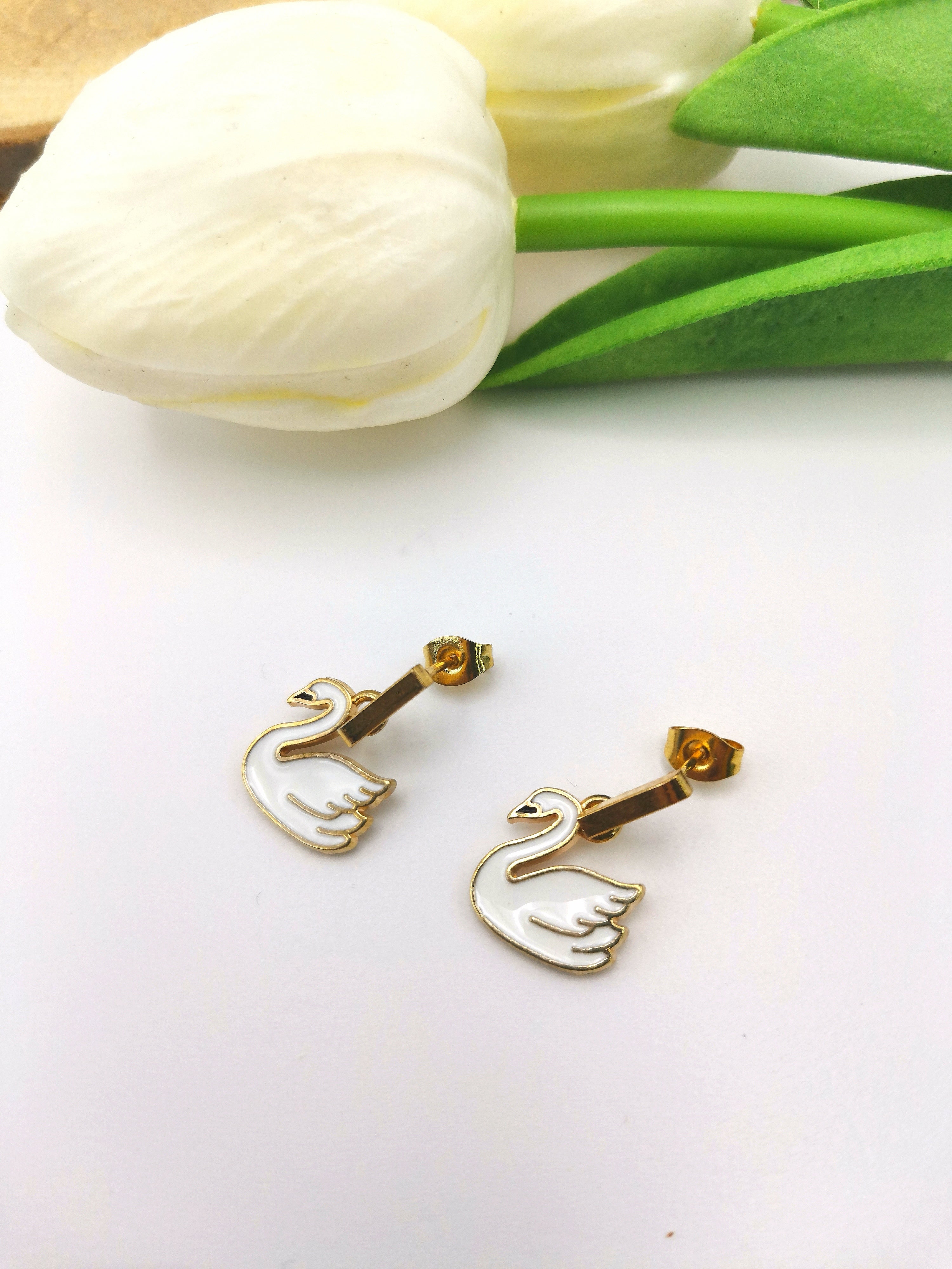 The Graceful Swan Earrings by Silver Sunbird, adorned with gold-plated enamel swan charms, are showcased on a white surface. Their elegant design is enhanced by two white tulips and green leaves in the background, adding to their sophisticated allure.