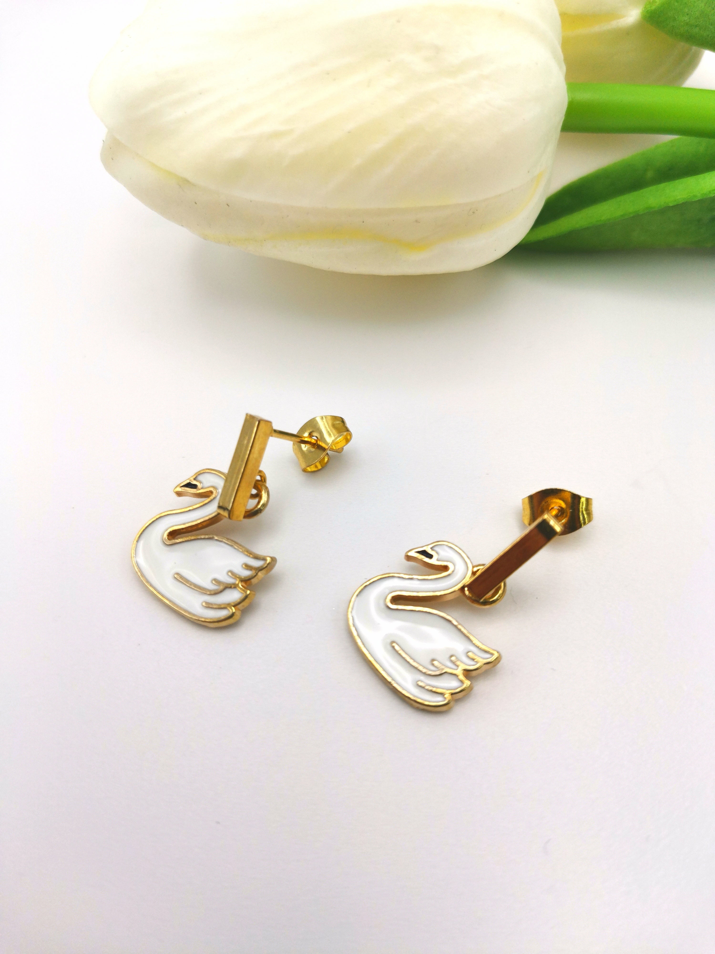 The Graceful Swan Earrings by Silver Sunbird feature gold-plated enamel swan charms, elegantly displayed on a white surface with a tulip backdrop. These handmade earrings exude sophistication and style, making them an ideal accessory for any occasion.