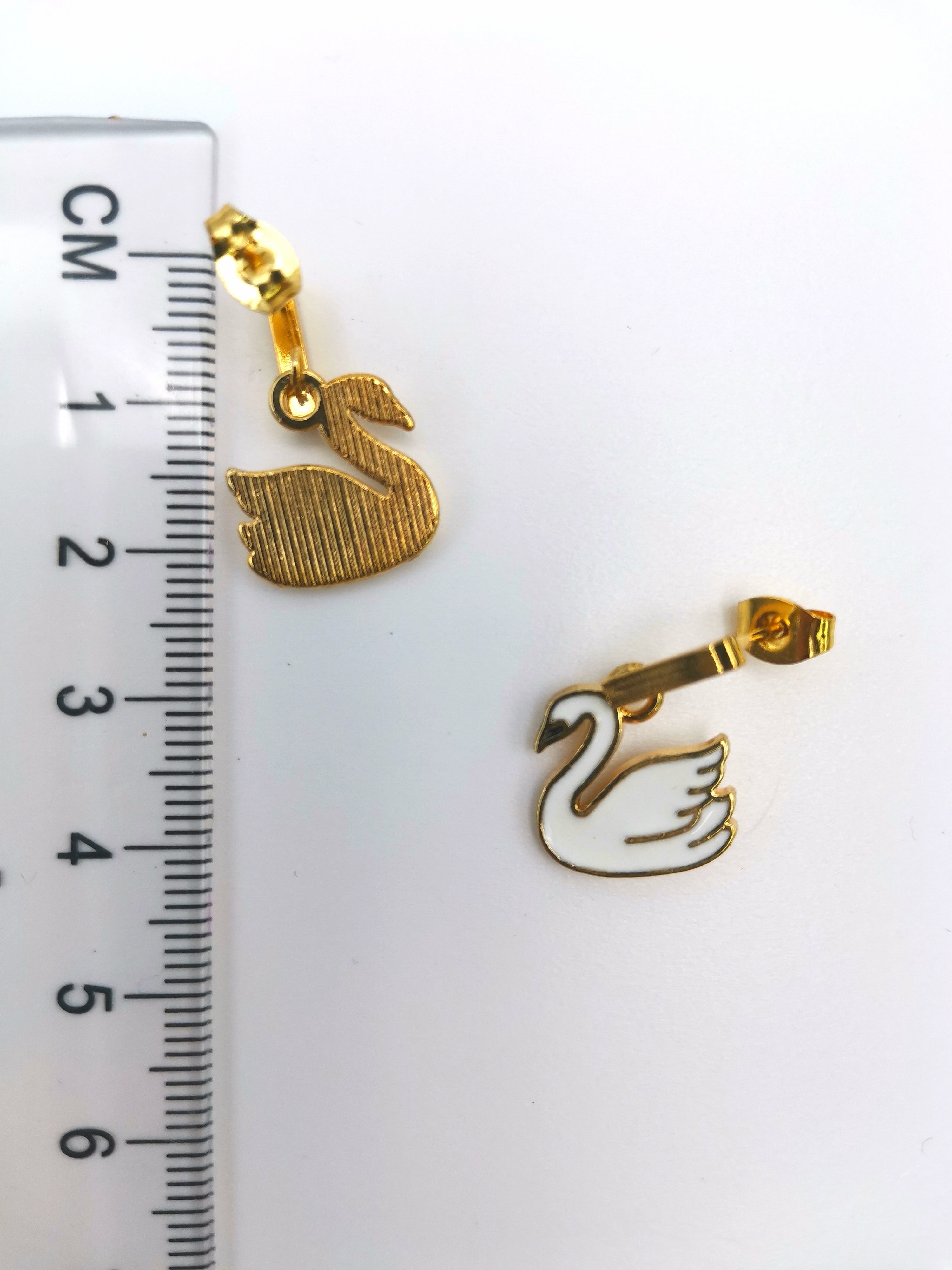 Graceful Swan Earrings by Silver Sunbird are pictured beside a ruler on a white background. One gold charm is textured, the other is smooth and outlined, ideal as elegant drop earrings or exquisite gold-plated enamel accents for handmade jewelry.