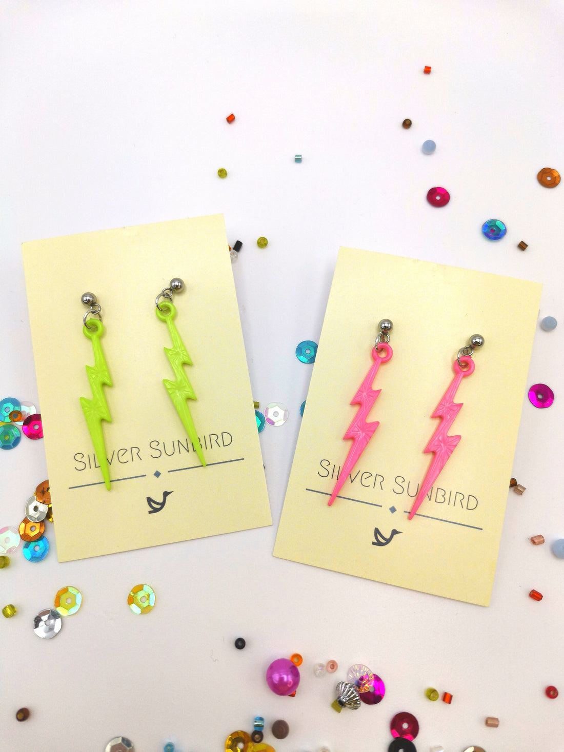 Two pairs of Electrifying Lightning Bolt Earrings from The Pastel Range by Silver Sunbird, one in neon green and the other in bright pink, crafted with hypoallergenic stainless steel, are showcased on Silver Sunbird backing cards with colorful sequins and small beads scattered around.