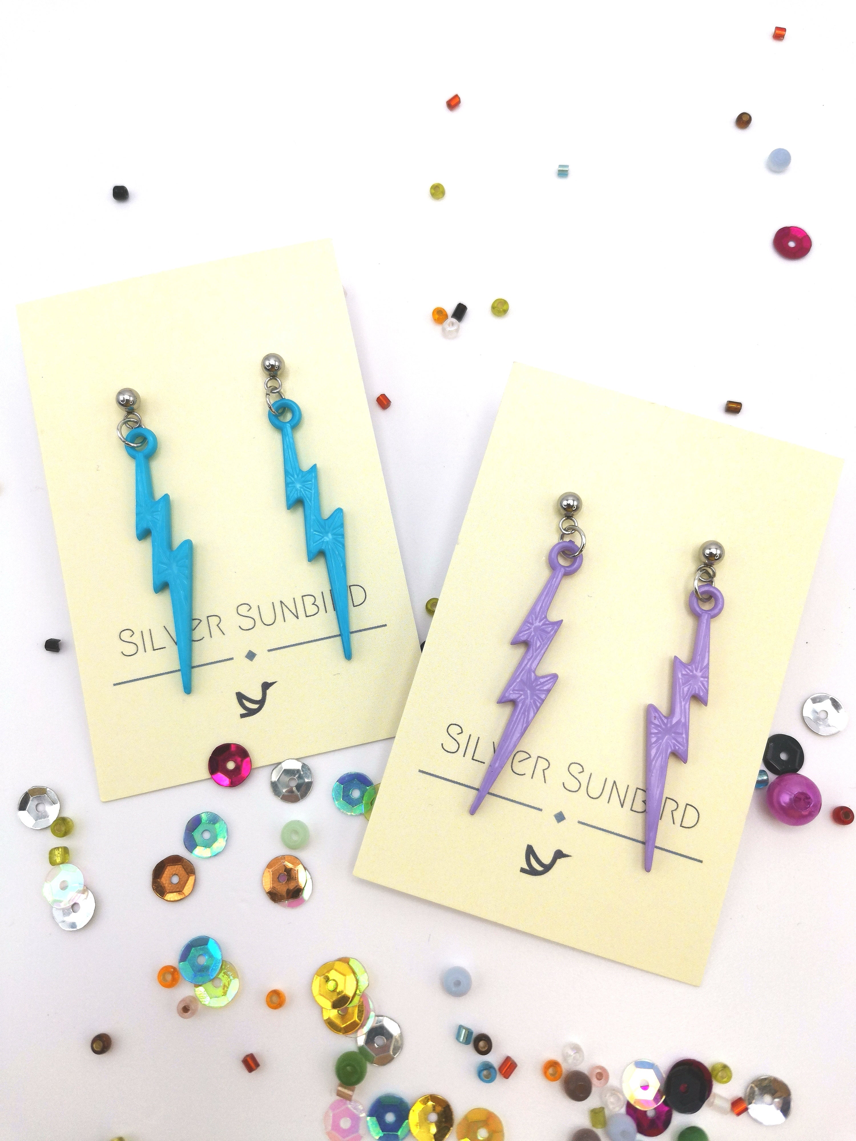 Two pairs of Electrifying Lightning Bolt Earrings from the Pastel Range by Silver Sunbird, crafted with hypoallergenic stainless steel. One pair is blue and the other purple, presented on cards with a colorful sequin-adorned background.