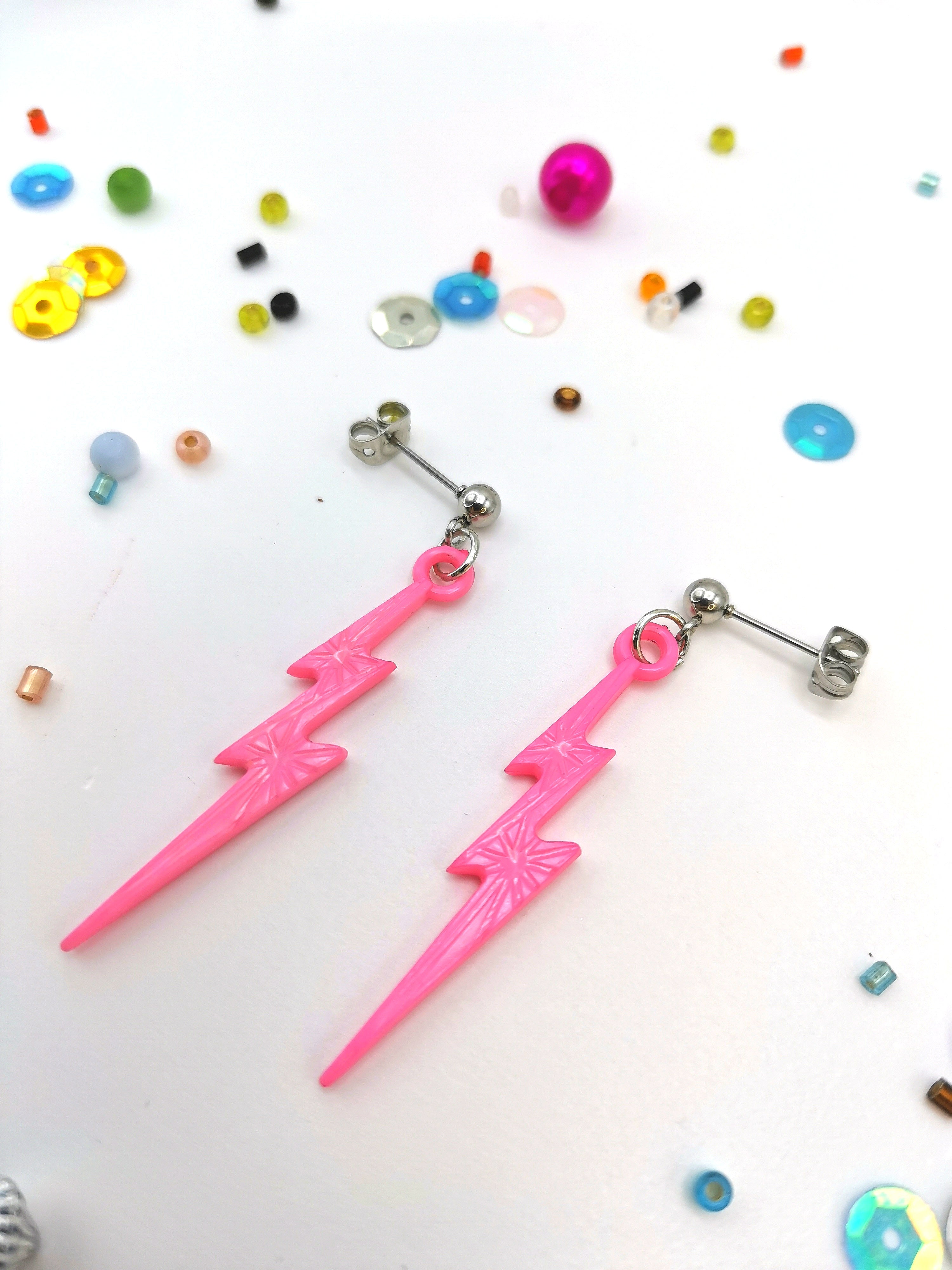 The Electrifying Lightning Bolt Earrings from The Pastel Range by Silver Sunbird feature hypoallergenic stainless steel studs, resting on a white surface amidst colorful beads and sequins.