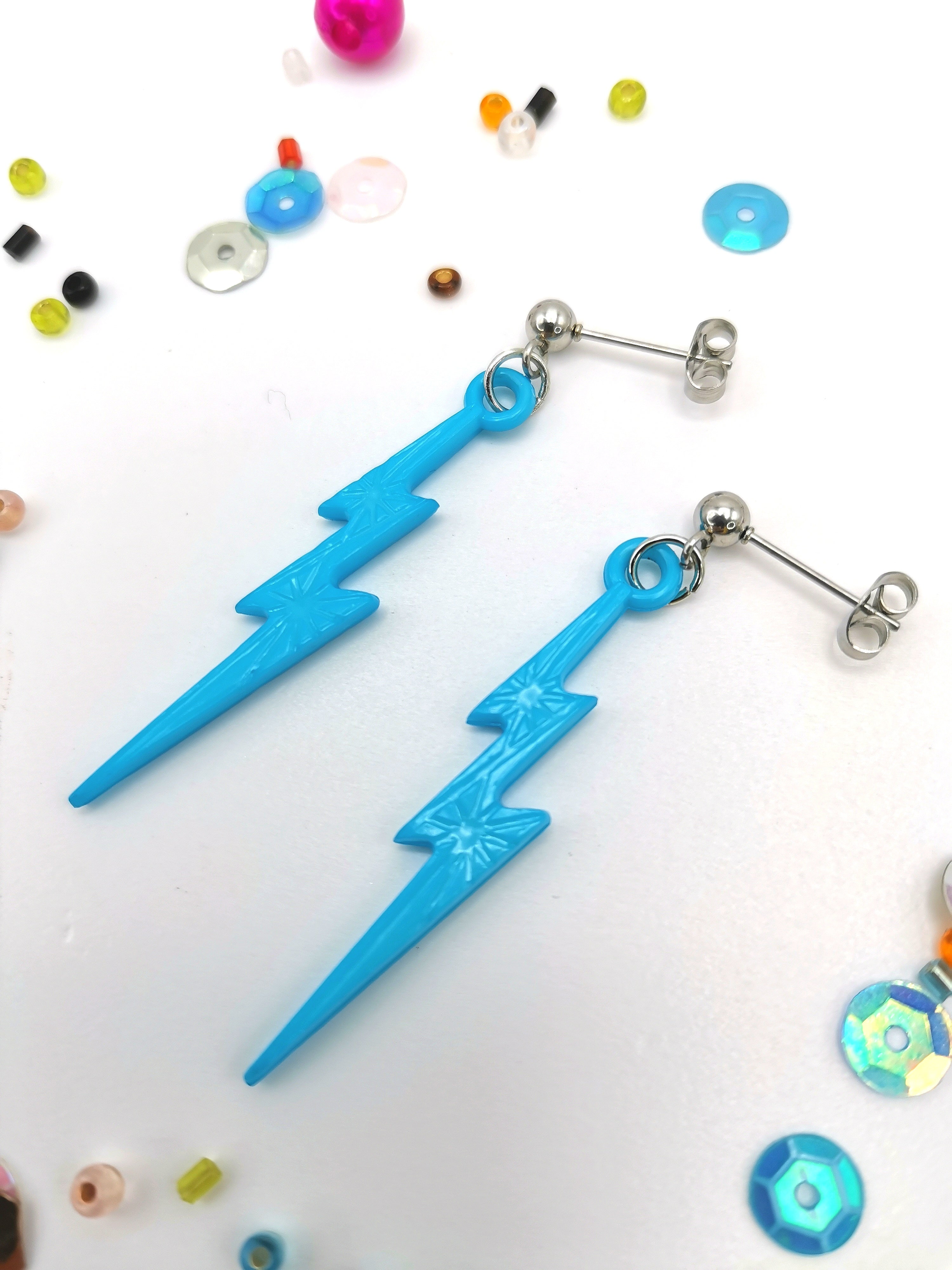 Two Electrifying Lightning Bolt Earrings from The Pastel Range by Silver Sunbird, with blue star patterns in hypoallergenic stainless steel, are adorned with colorful beads and sequins on a white background.