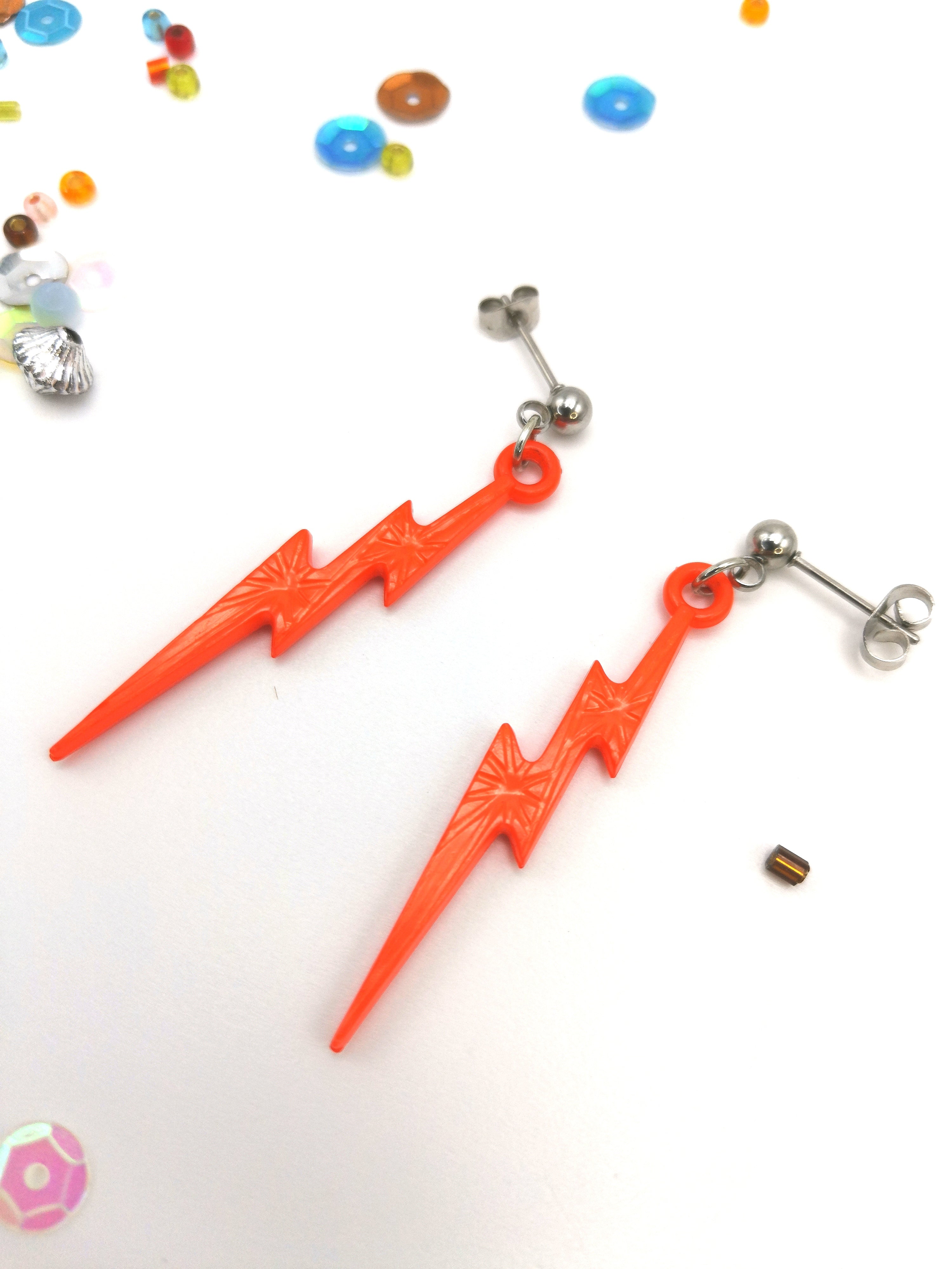 The Silver Sunbird Electrifying Lightning Bolt Earrings from The Bold Range feature bright orange lightning bolts with metallic studs on a white base, highlighted by colorful beads and shimmering sequins. Crafted from hypoallergenic stainless steel, they add a vibrant edge to any outfit.