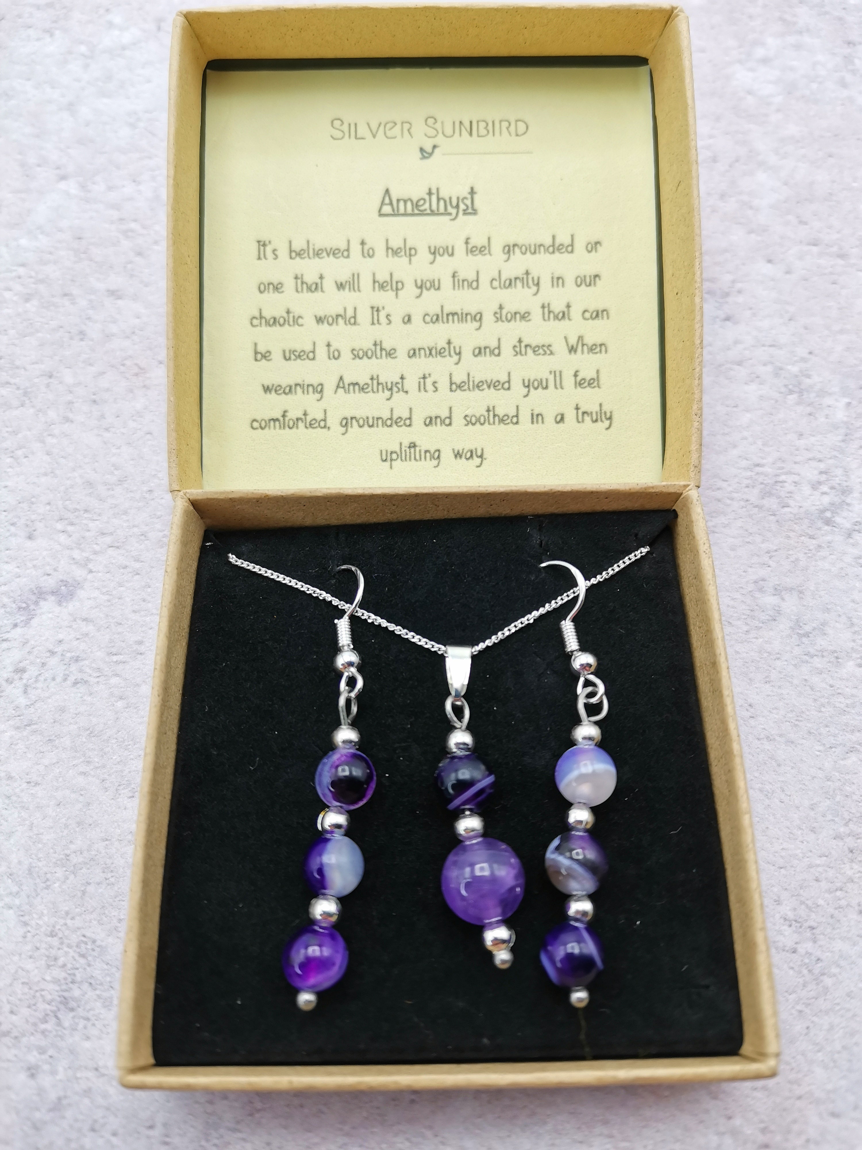 The Silver Sunbird Amethyst Gemstone Jewellery Set, featuring a necklace and matching earrings in 925 sterling silver, is arranged on black velvet in an open box. The lid highlights amethyst&