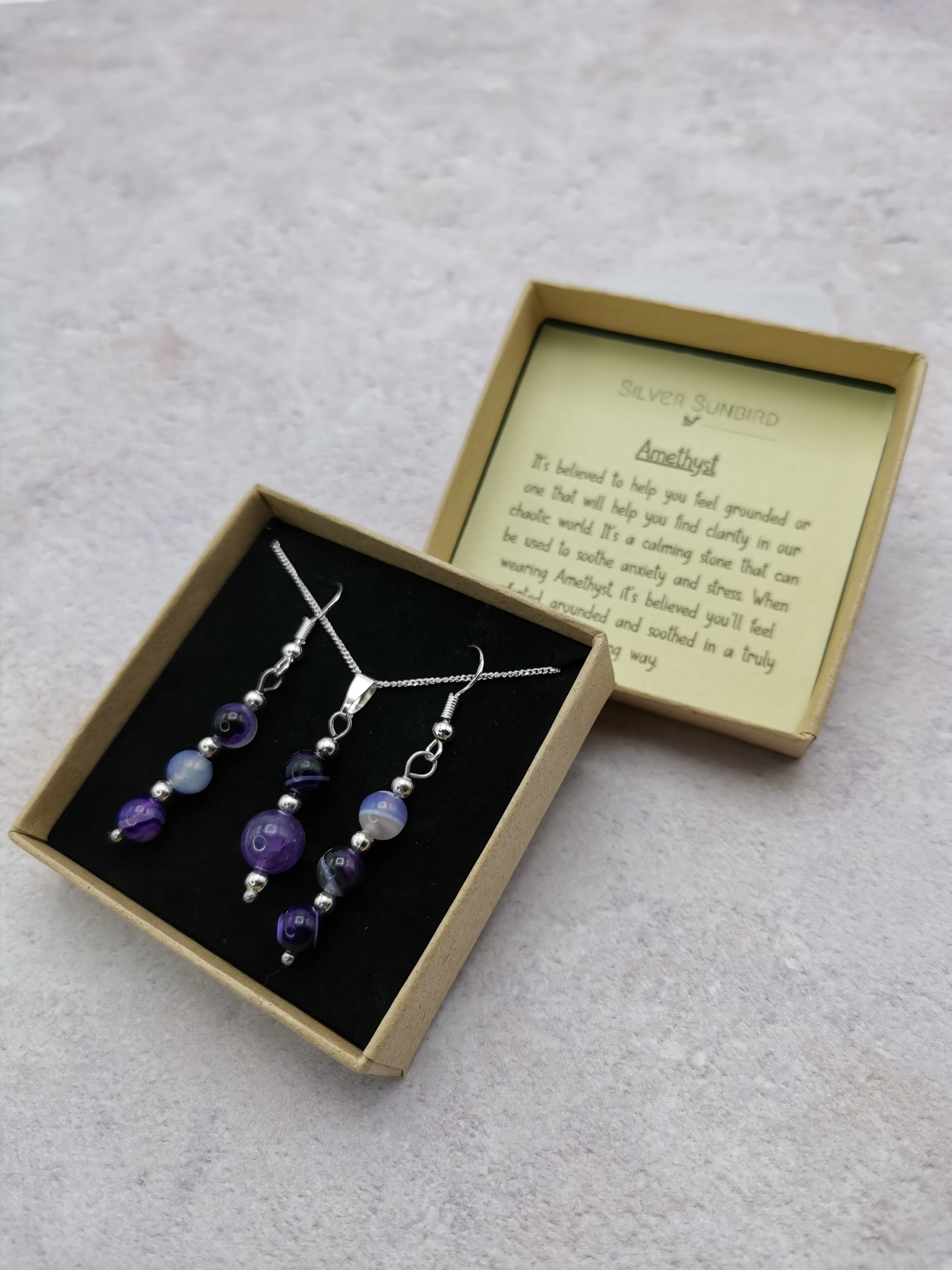 The Silver Sunbird Amethyst Gemstone Jewellery Set includes two 925 sterling silver necklaces with purple and blue pendants, elegantly presented in an open beige box. The lid highlights the amethyst&