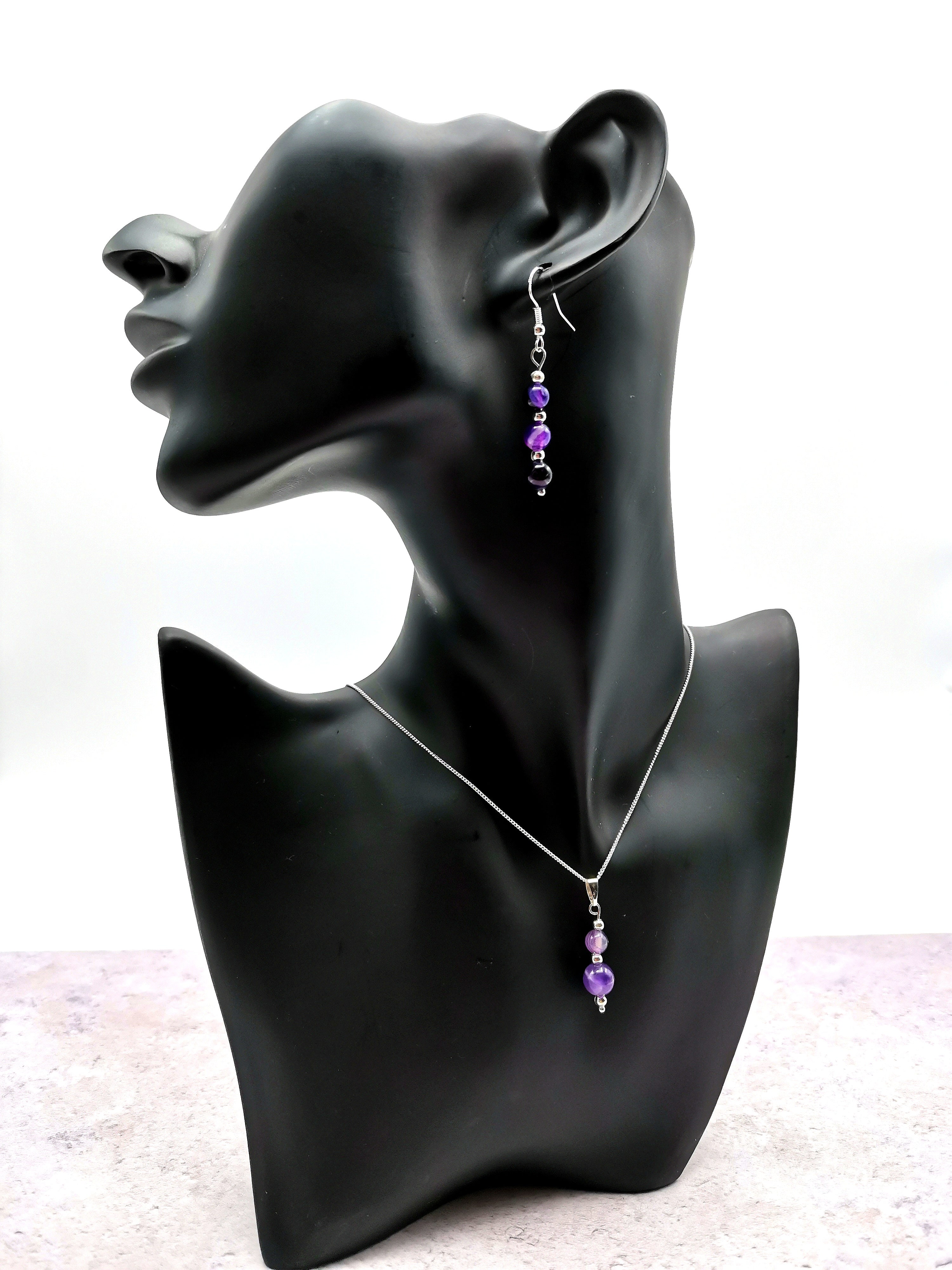 A black mannequin bust displays the Silver Sunbird Amethyst Gemstone Jewellery Set, featuring a silver necklace and matching earrings embellished with purple beads. The soft gray background enhances the elegance of this collection.
