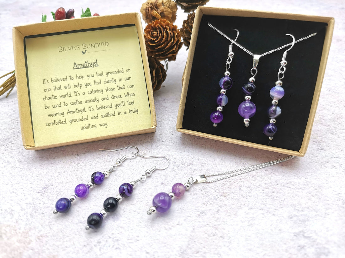 The Amethyst Gemstone Jewellery Set by Silver Sunbird, with shimmering purple beads adorning a necklace, bracelet, and earrings, is elegantly displayed in a black 925 sterling silver box. A card highlights the gemstone&