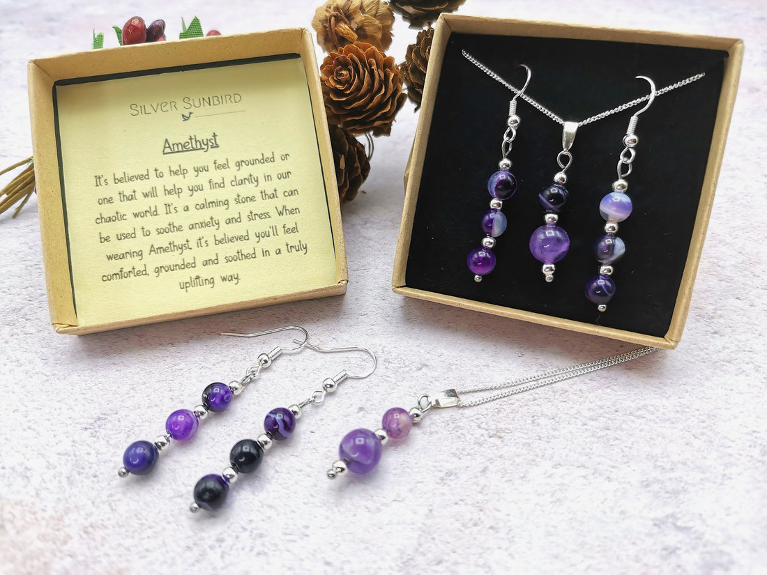The Silver Sunbird Amethyst Gemstone Jewellery Set, with purple beads and 925 sterling silver, includes a necklace, bracelet, and earrings in an open box. A card details its calming properties against a backdrop of pinecones.