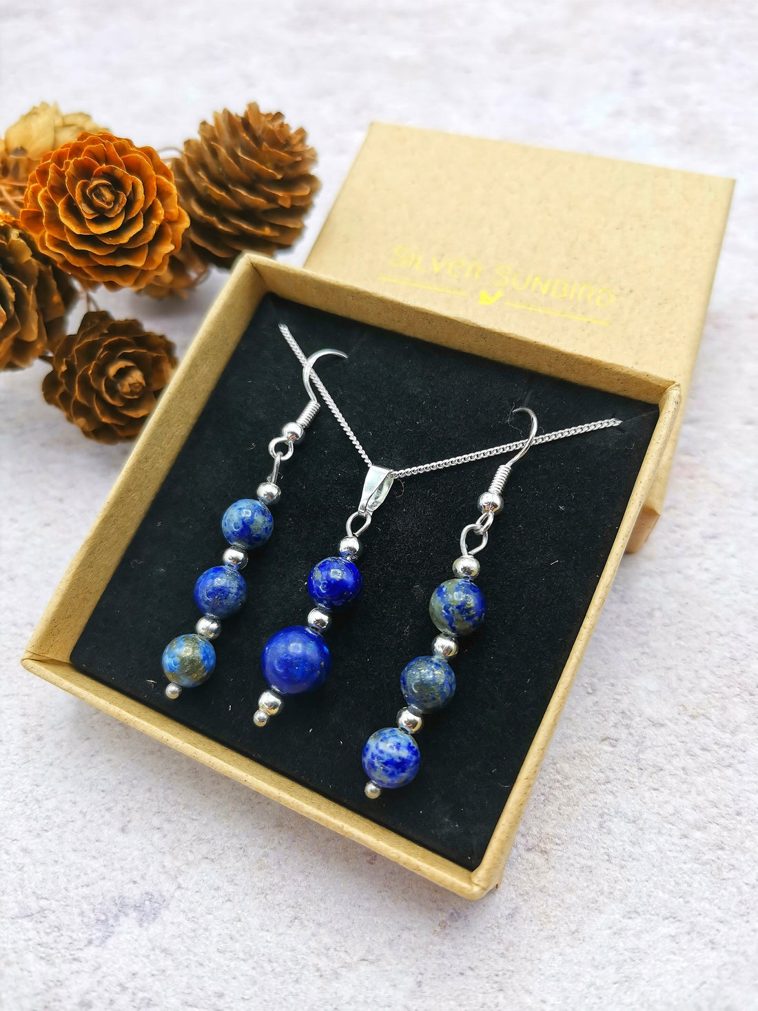 The Silver Sunbird Lapis Lazuli Gemstone Jewellery Set arrives in a beige box, featuring a necklace and earrings with lapis lazuli-like blue beads and 925 sterling silver spacers. Pine cones next to the box add a natural touch to the light-colored surface.