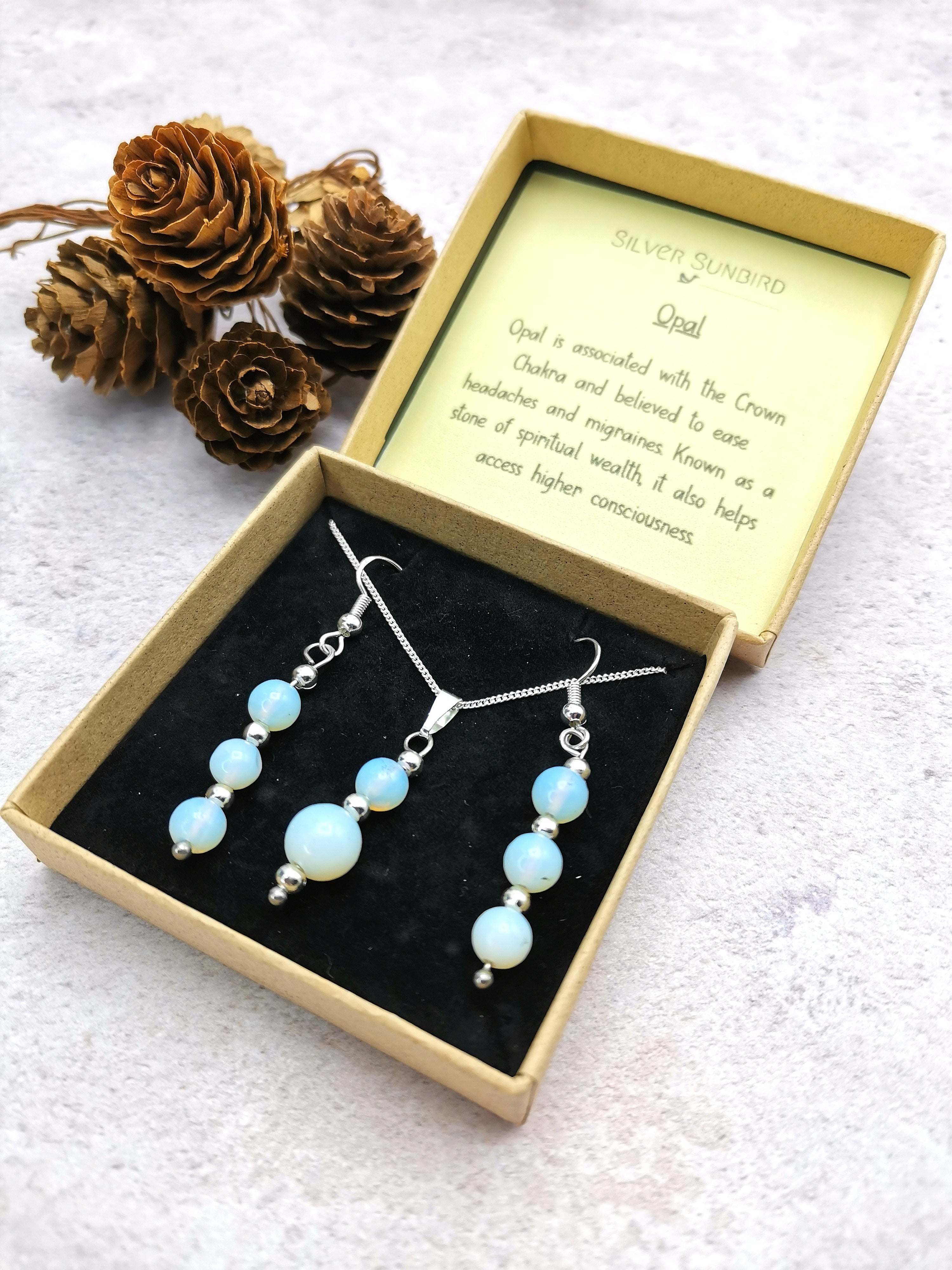 The Silver Sunbird Opal Gemstone Gift Set, featuring opal earrings and a necklace in sterling silver, is elegantly displayed in an open box. A card explaining opal&