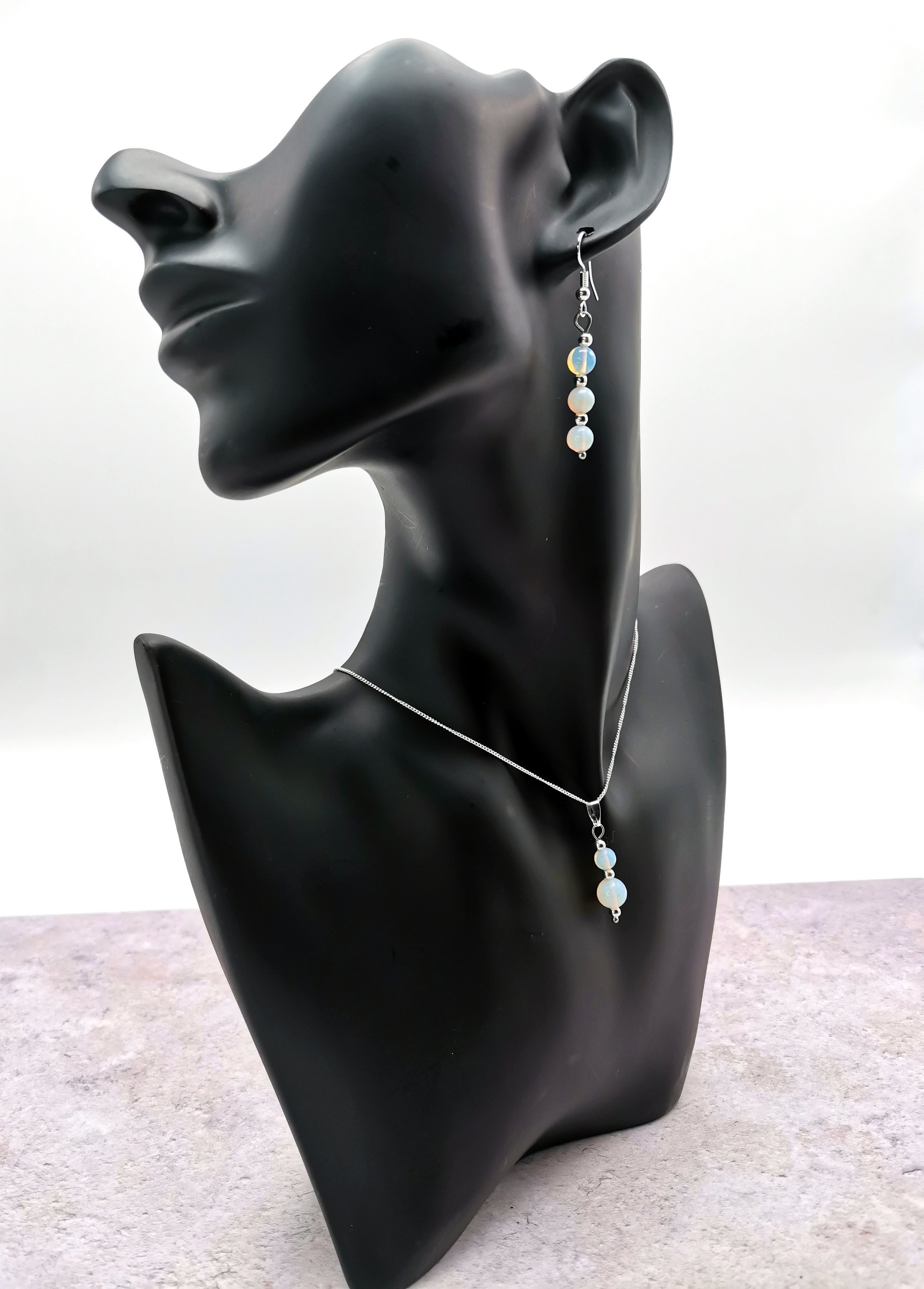 The Silver Sunbird Opal Gemstone Gift Set, featuring opal earrings and a matching necklace with light blue and pale yellow beads set in sterling silver, is elegantly showcased on a black mannequin bust against a neutral background.