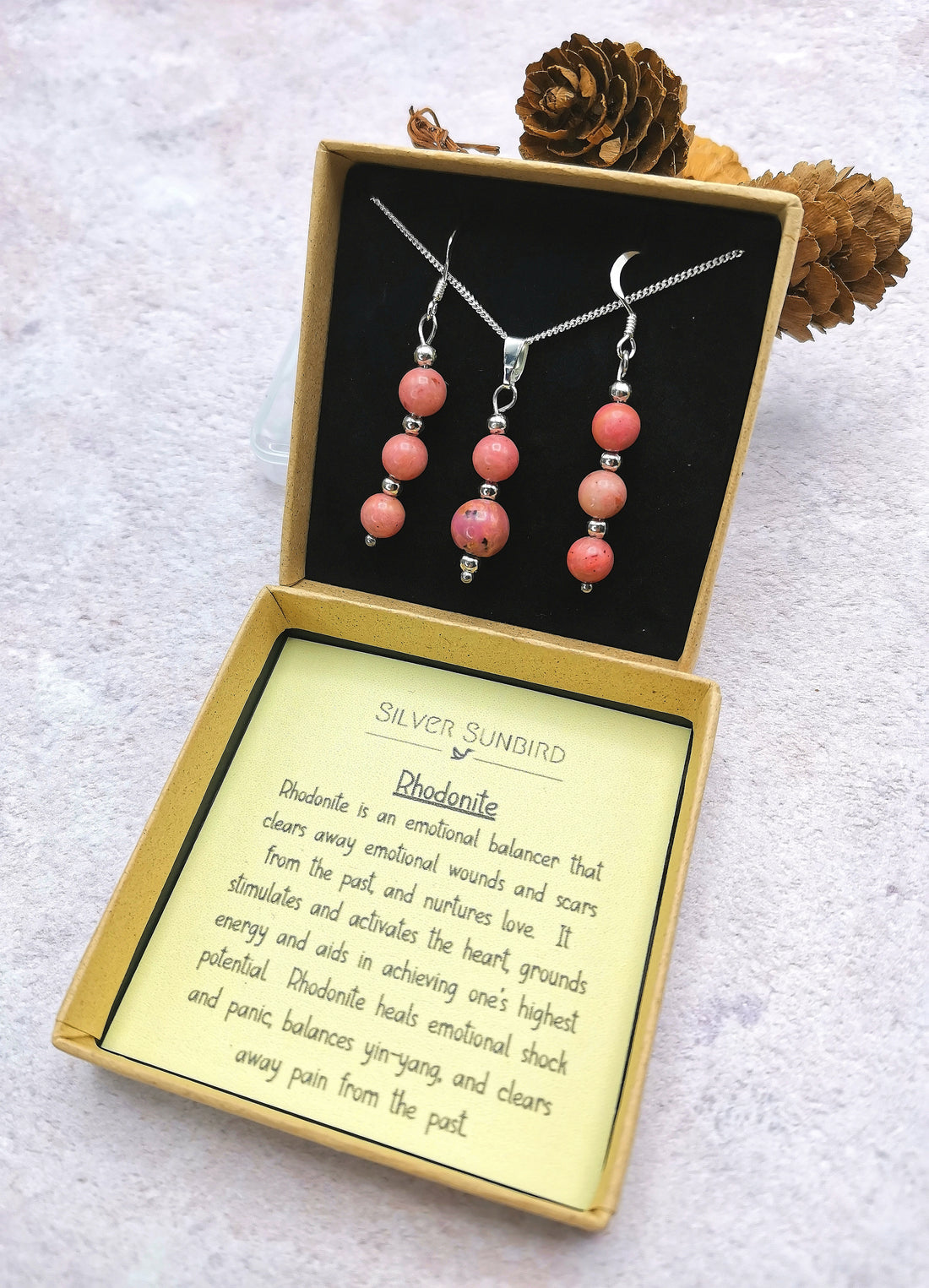The Silver Sunbird Rhodonite Gemstone Jewellery Set includes a necklace and earrings in a box with details on rhodonite’s emotional healing properties. Featuring sterling silver accents, it&