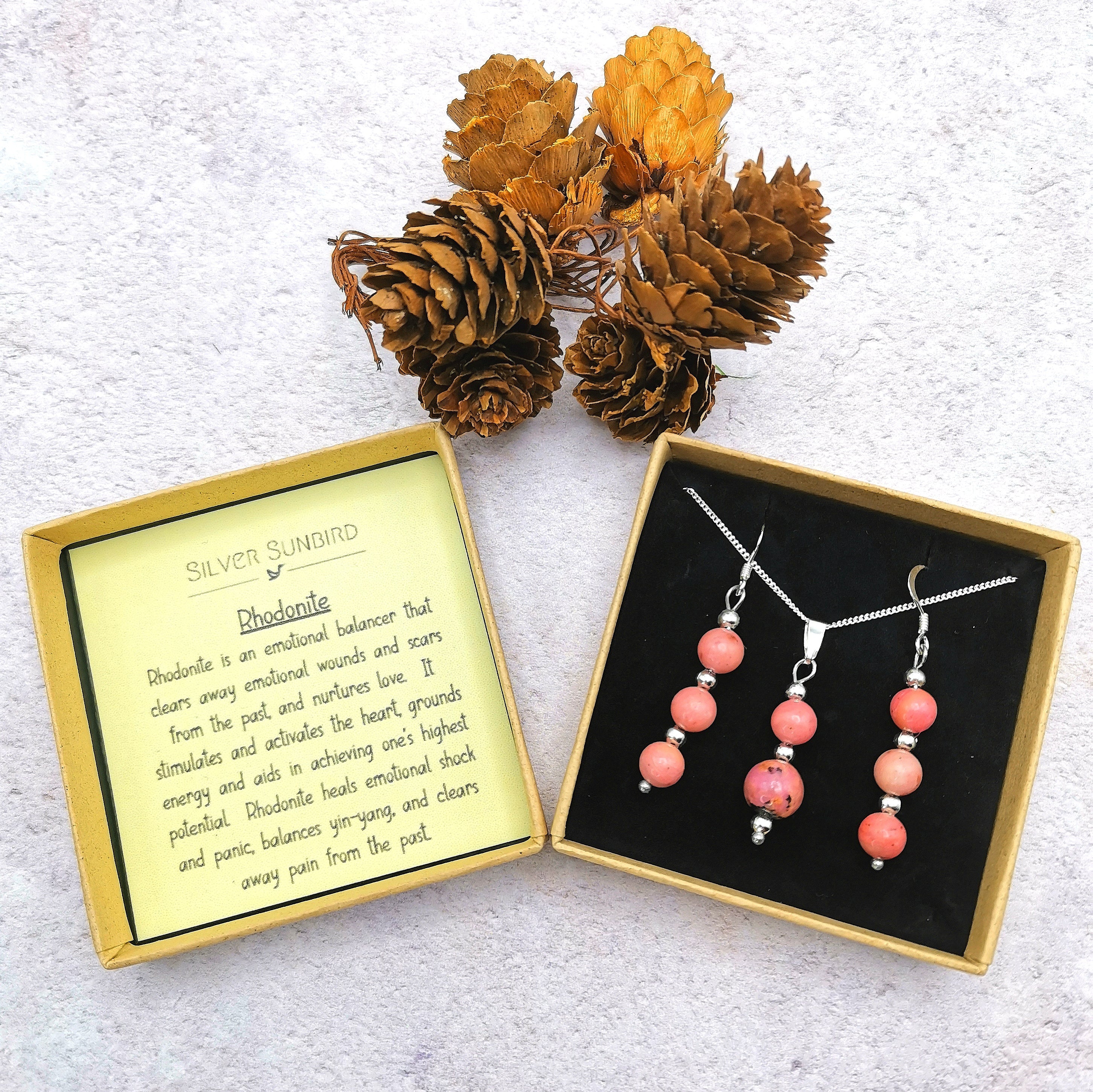 Two open Silver Sunbird jewelry boxes sit on a white surface: one with pink bead earrings and a matching Rhodonite Gemstone necklace, and the other with a card detailing rhodonite&