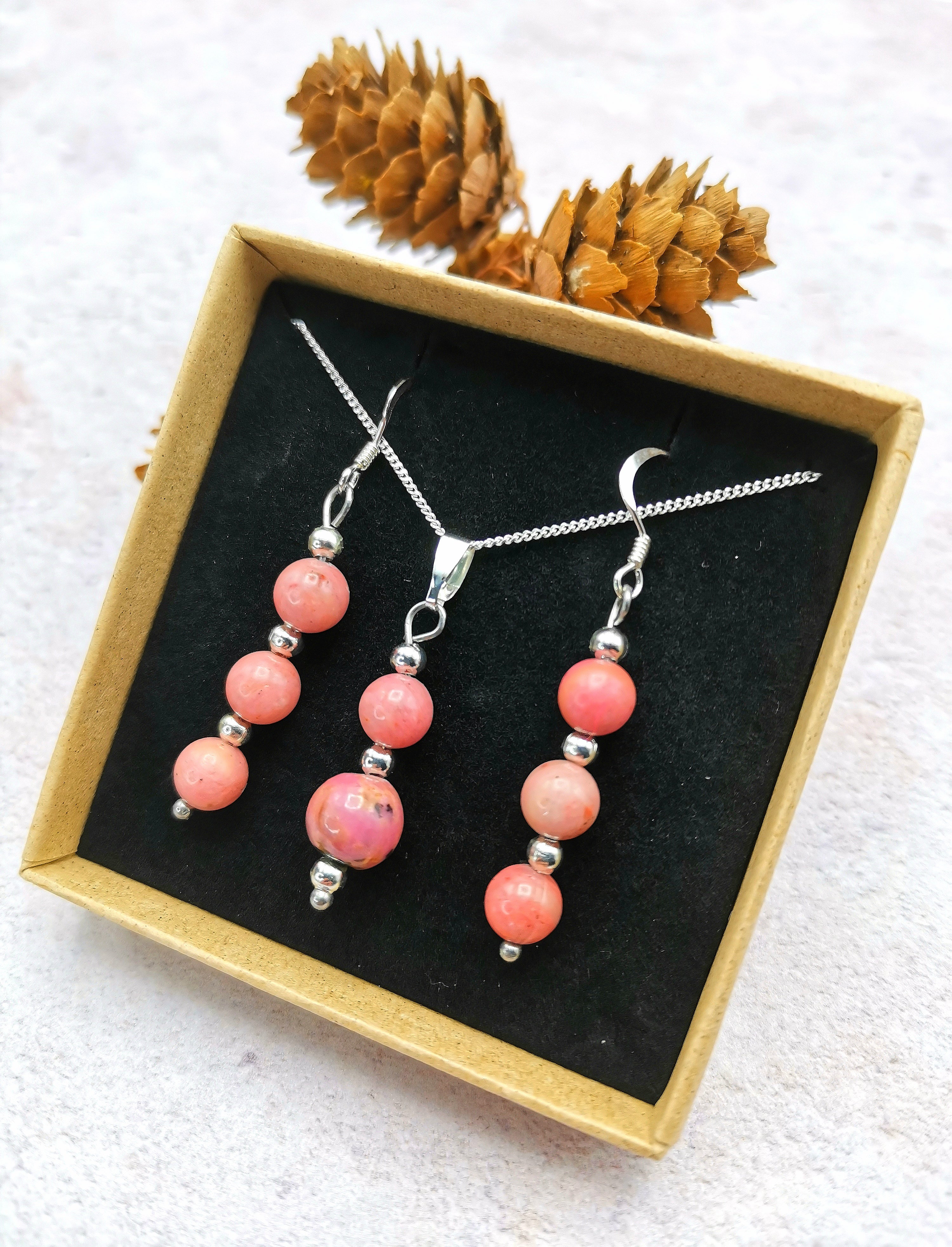 The Silver Sunbird Rhodonite Gemstone Jewellery Set includes a sterling silver necklace and earrings, beautifully presented in a box. Each piece features pink beads similar to Rhodonite gemstones with silver accents, set against an artful backdrop of pine cones on a textured surface.