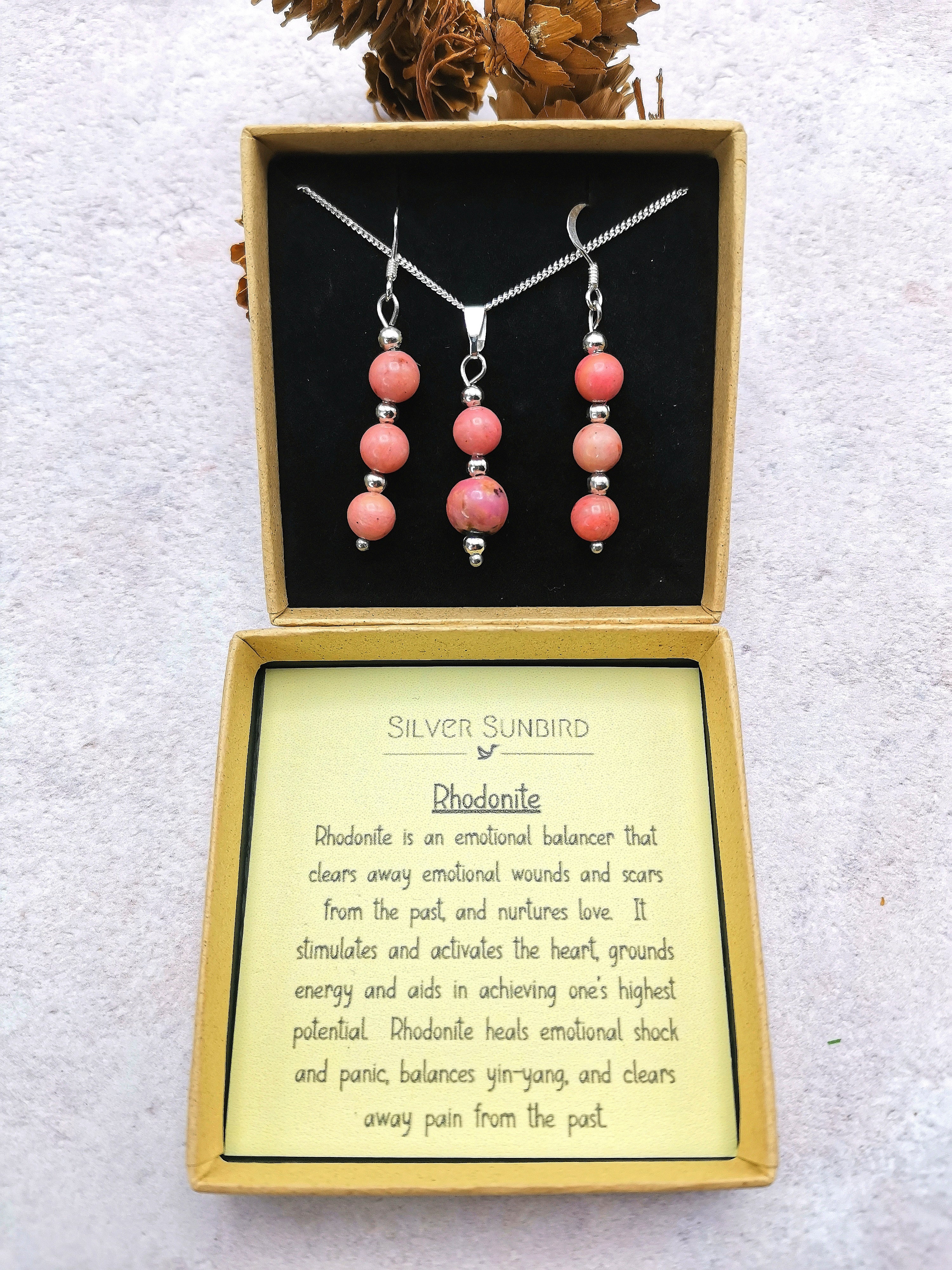 The Rhodonite Gemstone Jewellery Set by Silver Sunbird includes a sterling silver necklace and earrings with pink rhodonite beads, stylishly presented in a yellow box with a card detailing the stone&