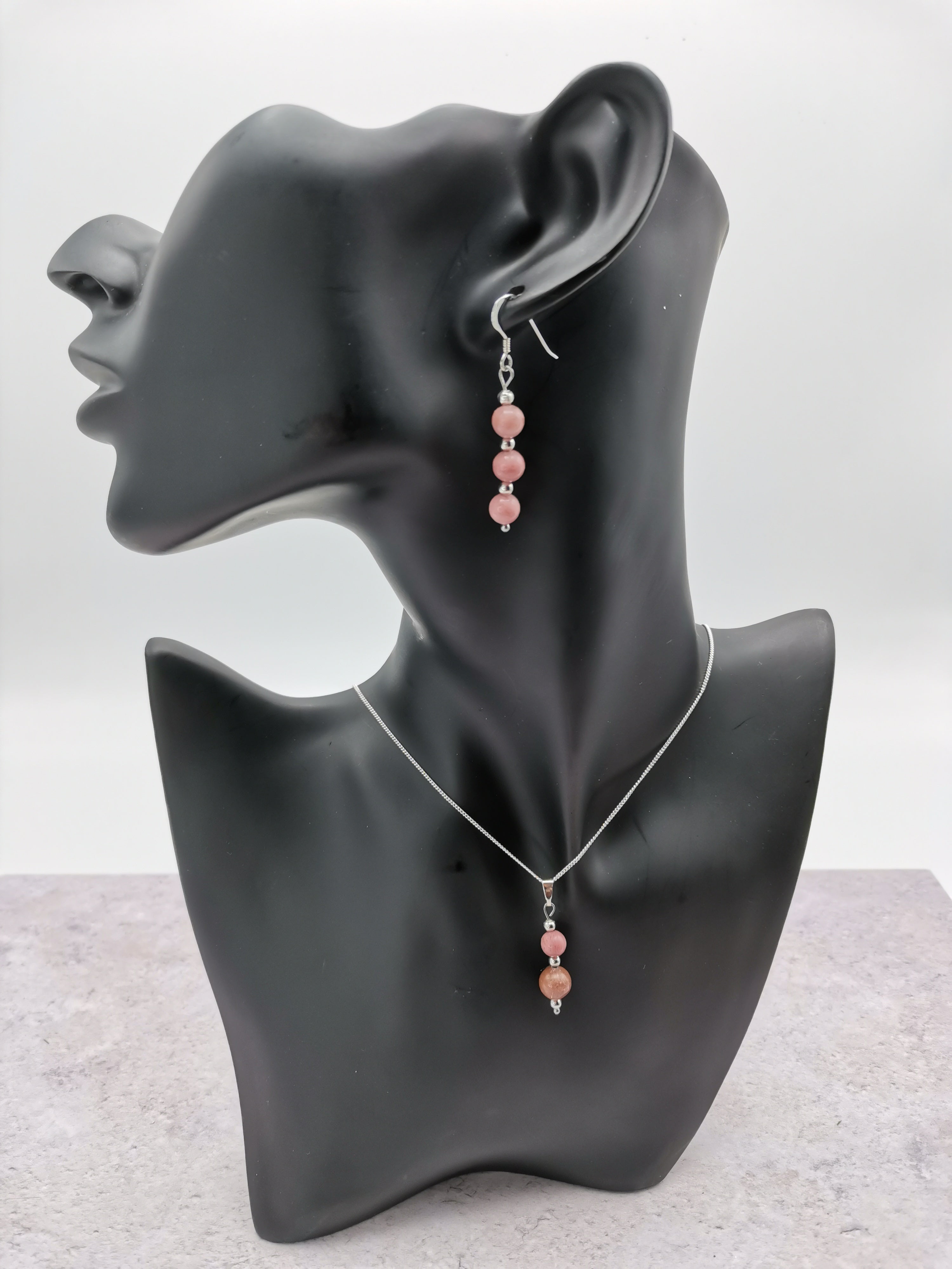 A black mannequin bust displays the Silver Sunbird Rhodonite Gemstone Jewellery Set, featuring sterling silver with pink beads, dangling earrings, and a matching necklace. The simple white and gray background accentuates its elegance.