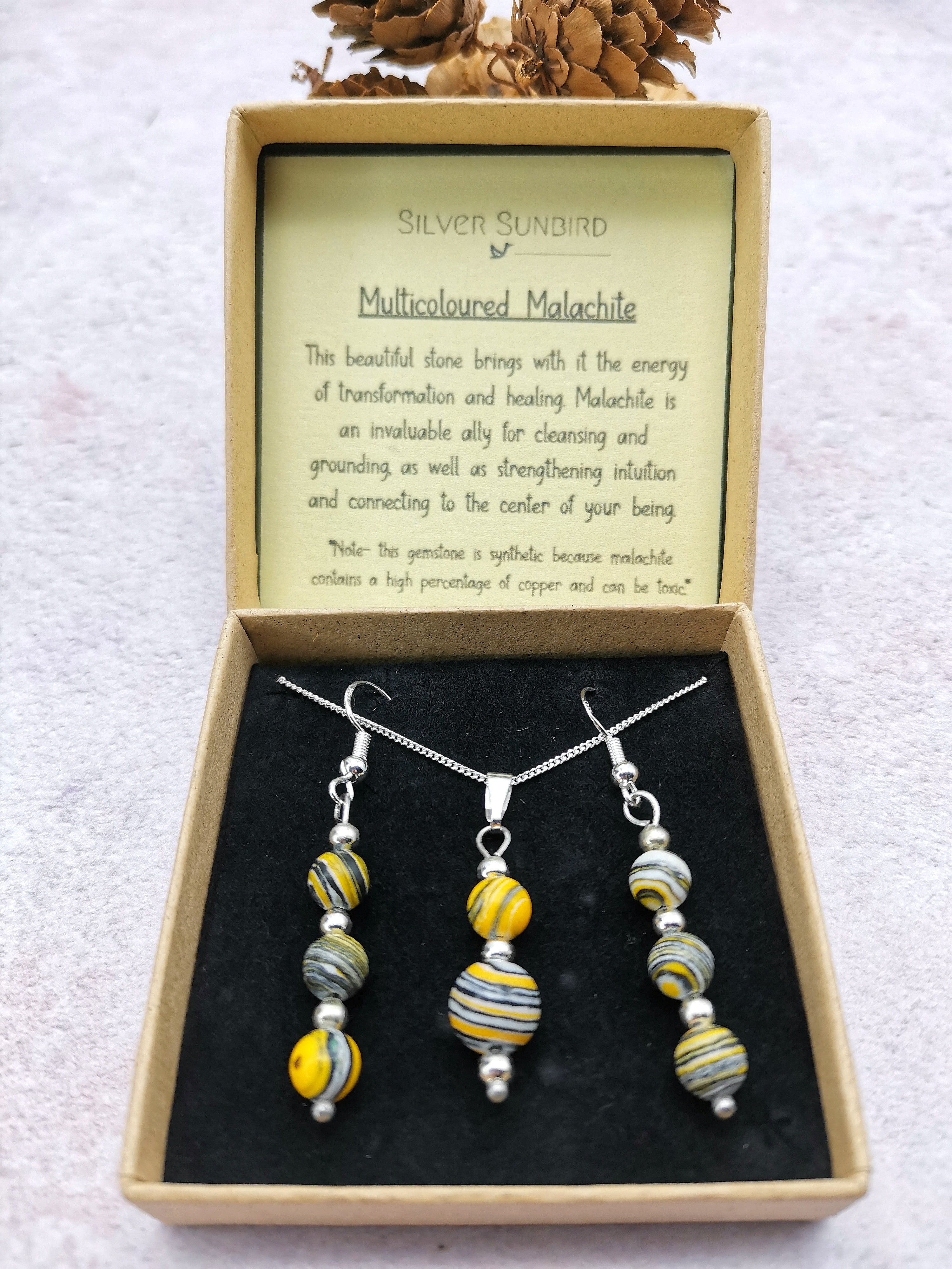 The Silver Sunbird "Multi-coloured Malachite Gemstone Gift Set" features yellow and gray striped beads in a light pink box with pinecones, highlighting malachite benefits and timeless elegance.