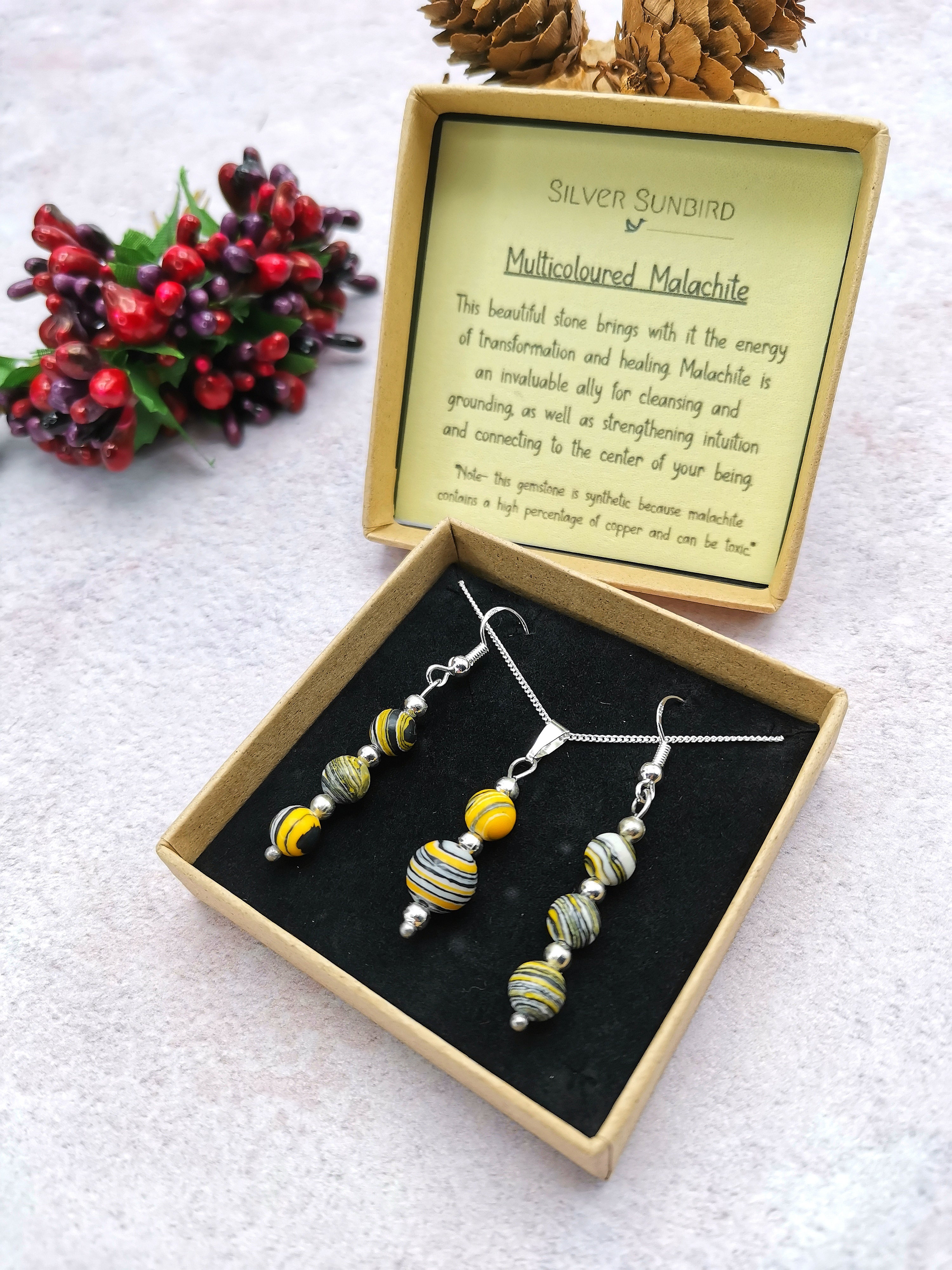 The Silver Sunbird Multi-coloured Malachite Gemstone Gift Set, featuring a sterling silver necklace and earrings with vibrant malachite beads, is elegantly presented in an open gift box complete with a malachite description. Pinecones and berries are artfully arranged in the background.