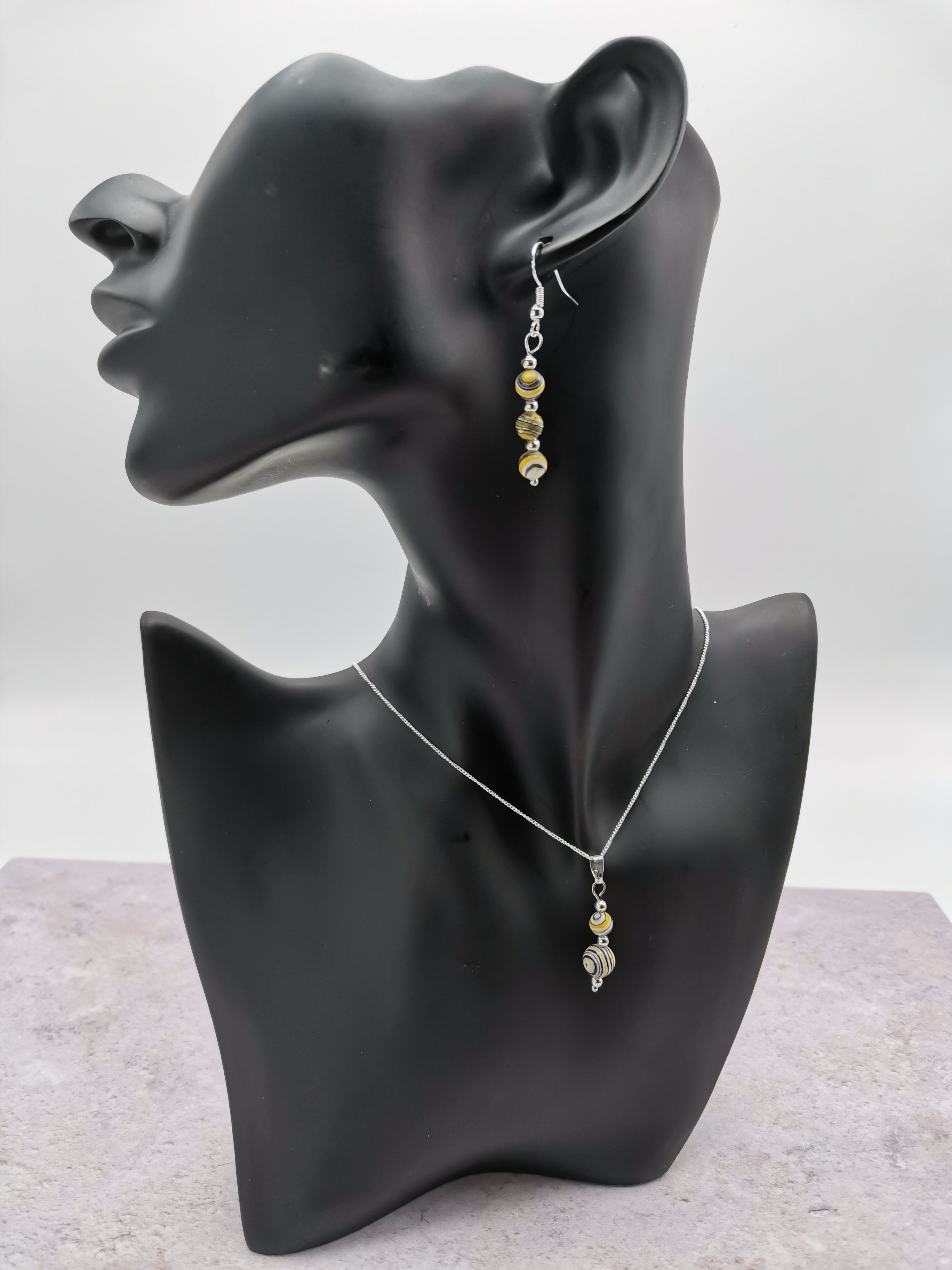 The Silver Sunbird Multi-coloured Malachite Gemstone Gift Set, displayed on a black mannequin bust against a soft gray backdrop, features stunning dangling earrings adorned with gold and silver beads, complemented by a sterling silver chain and pendant.