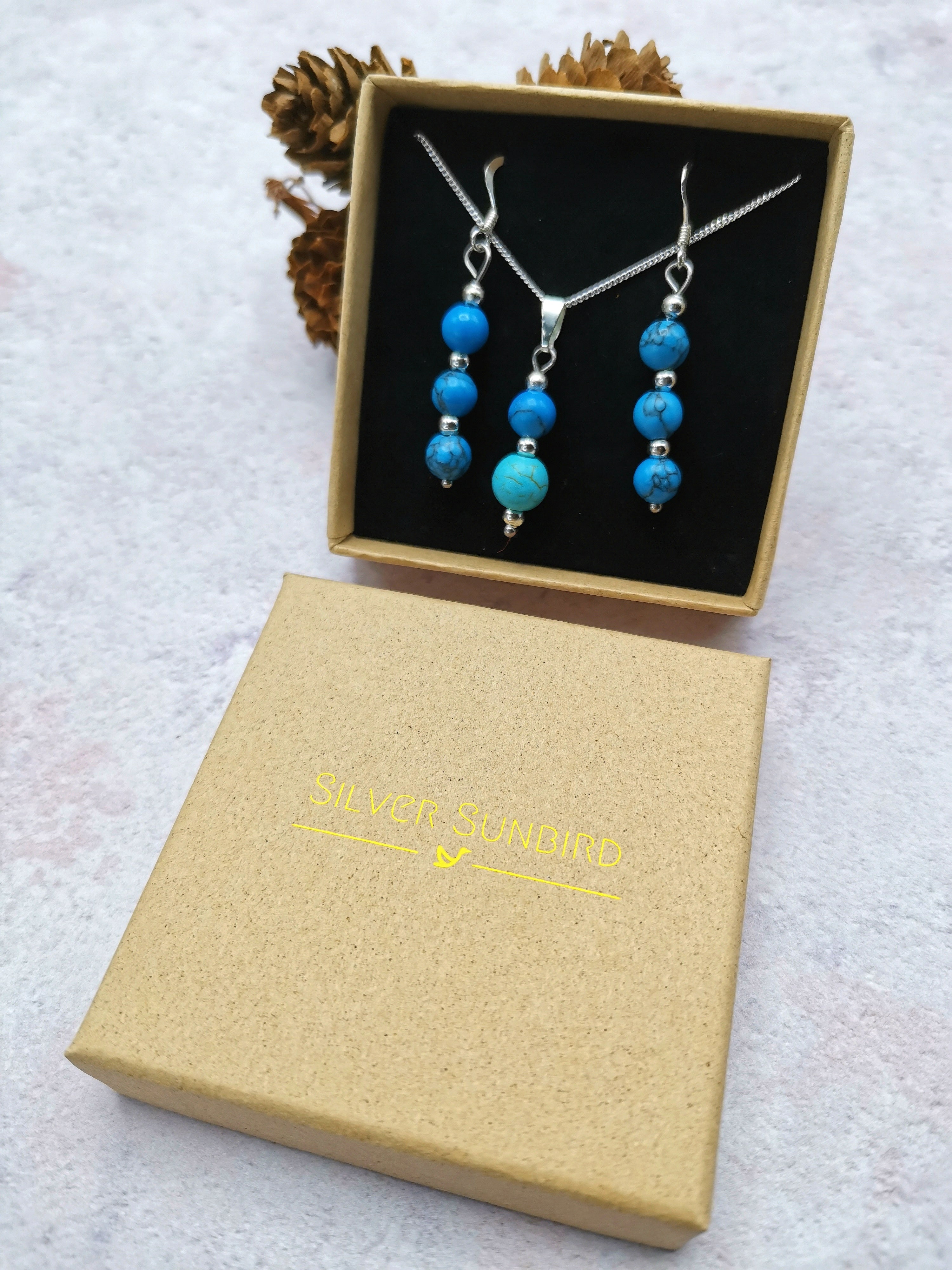 The Silver Sunbird Turquoise Gemstone Jewellery Set features a sterling silver necklace and matching earrings in a black velvet-lined brown box. A separate closed box labeled "SILVER SUNBIRD" is shown with pine cones in the background.