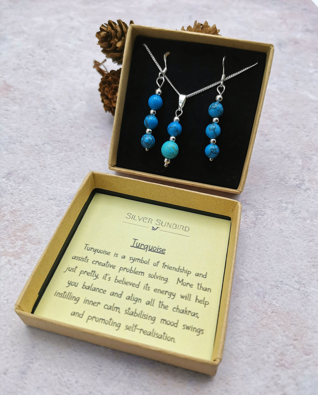 The Silver Sunbird Turquoise Gemstone Jewellery Set—featuring a sterling silver necklace and earrings—is displayed in an open box, with the lid highlighting turquoise’s symbolism of friendship, balance, and self-realisation. Pinecones accent the background.