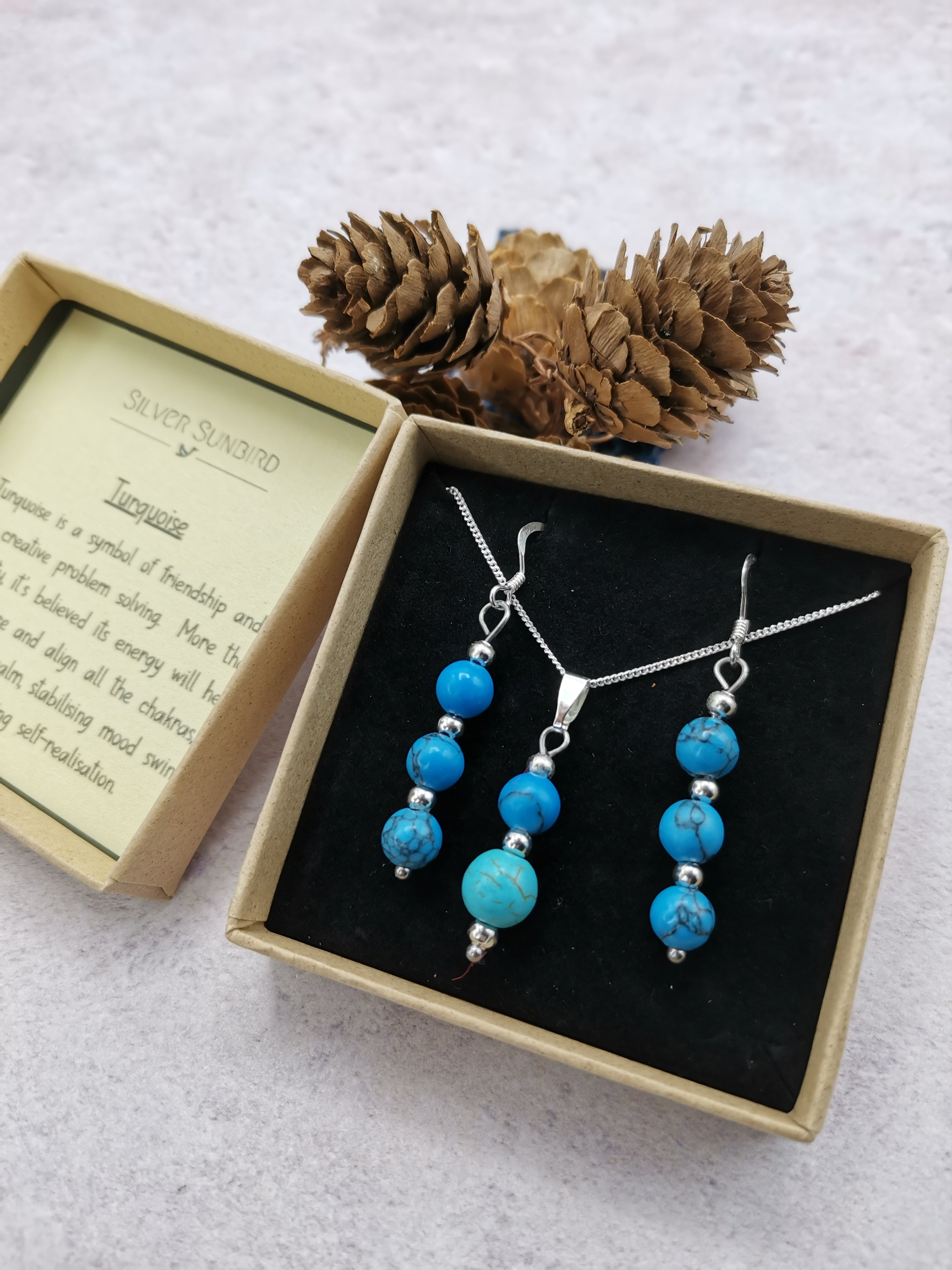 The Silver Sunbird Turquoise Gemstone Jewellery Set includes a sterling silver necklace and matching earrings, presented in a gift box with an info card and pine cone on a light textured surface.