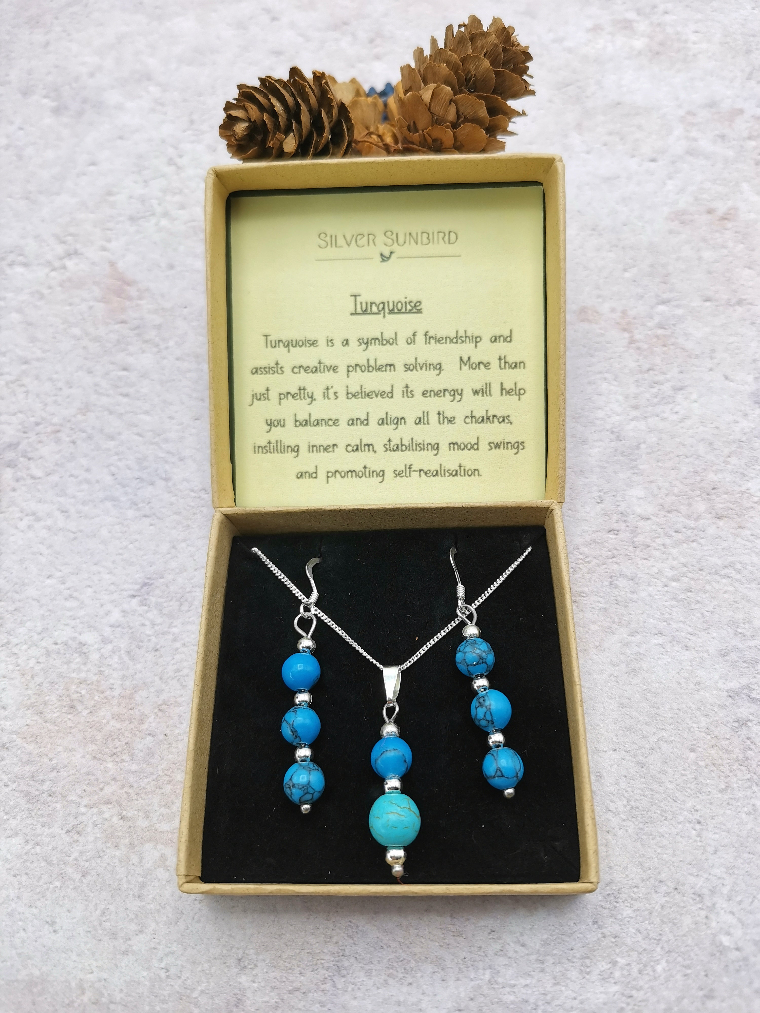 The Silver Sunbird Turquoise Gemstone Jewellery Set—featuring sterling silver necklace and earrings—is displayed in an open box, with a card on turquoise’s meaning inside the lid and two pine cones placed on top.