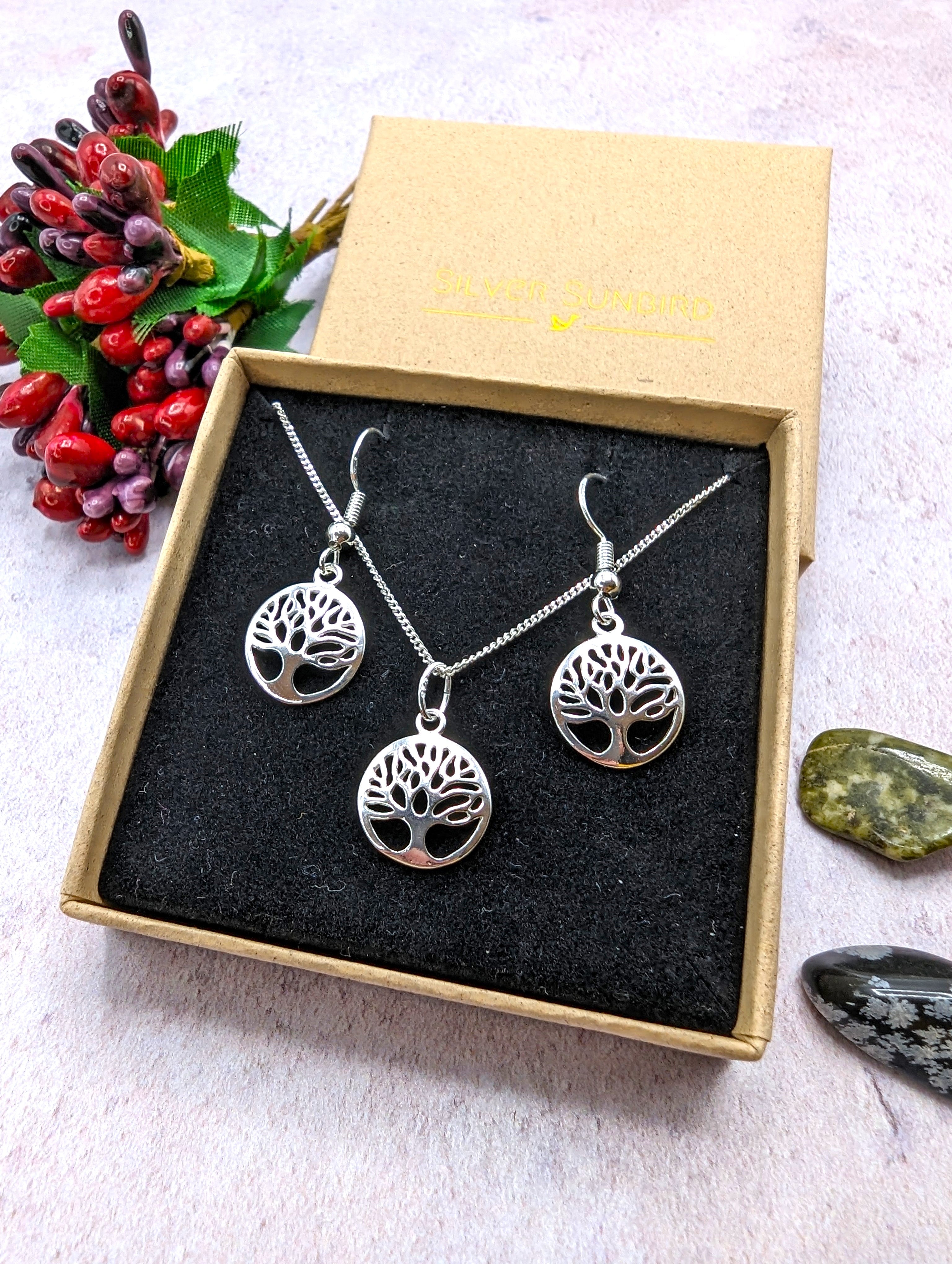 The Silver Sunbird Sterling Silver Tree of Life Jewellery Set includes a necklace and hypoallergenic earrings in a brown box with black velvet lining, accented by red berries and polished stones beside it.