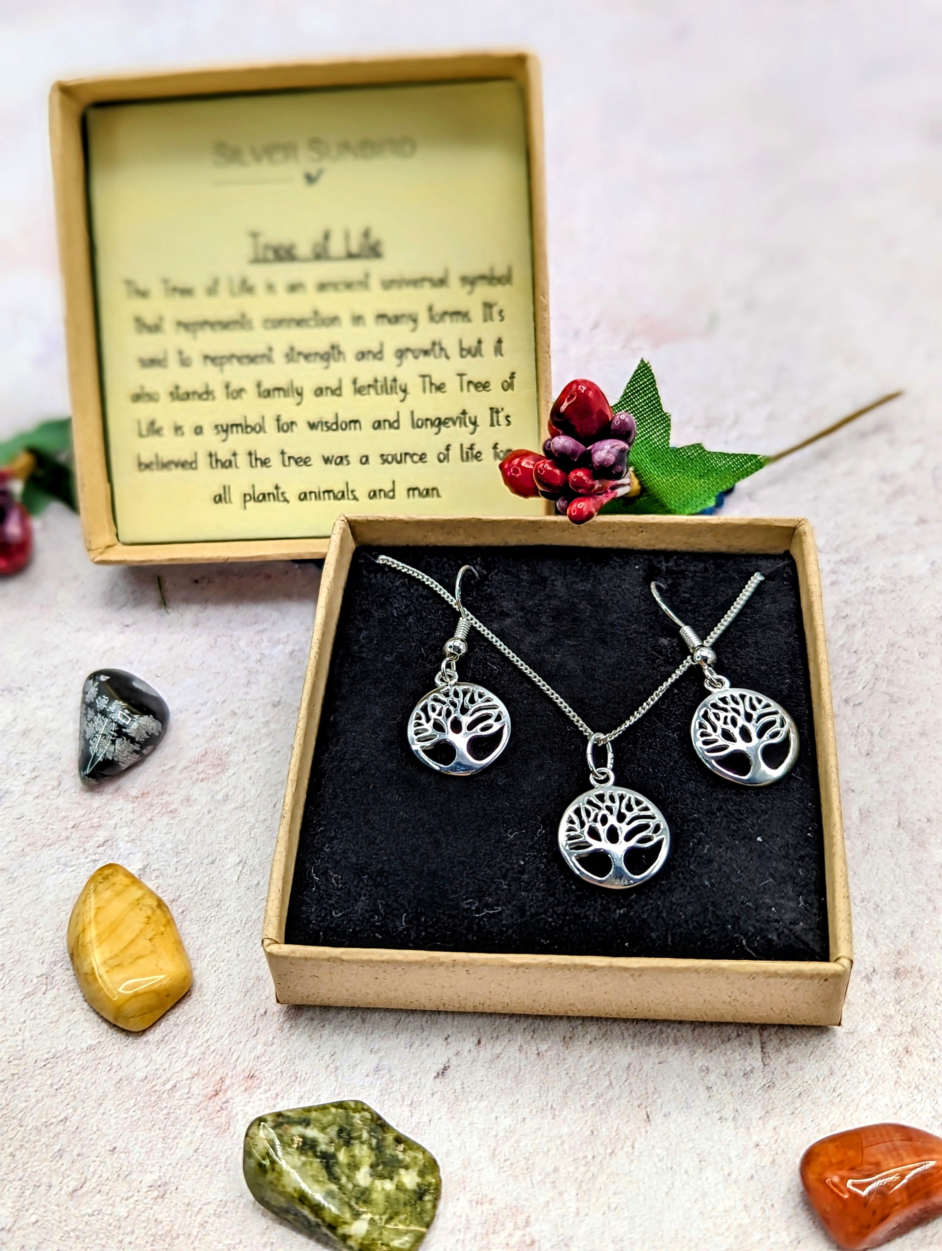 The Silver Sunbird Sterling Silver Tree of Life Jewellery Set includes a necklace and hypoallergenic earrings, beautifully displayed in a brown box with a description card, colorful stones, and a decorative berry branch.