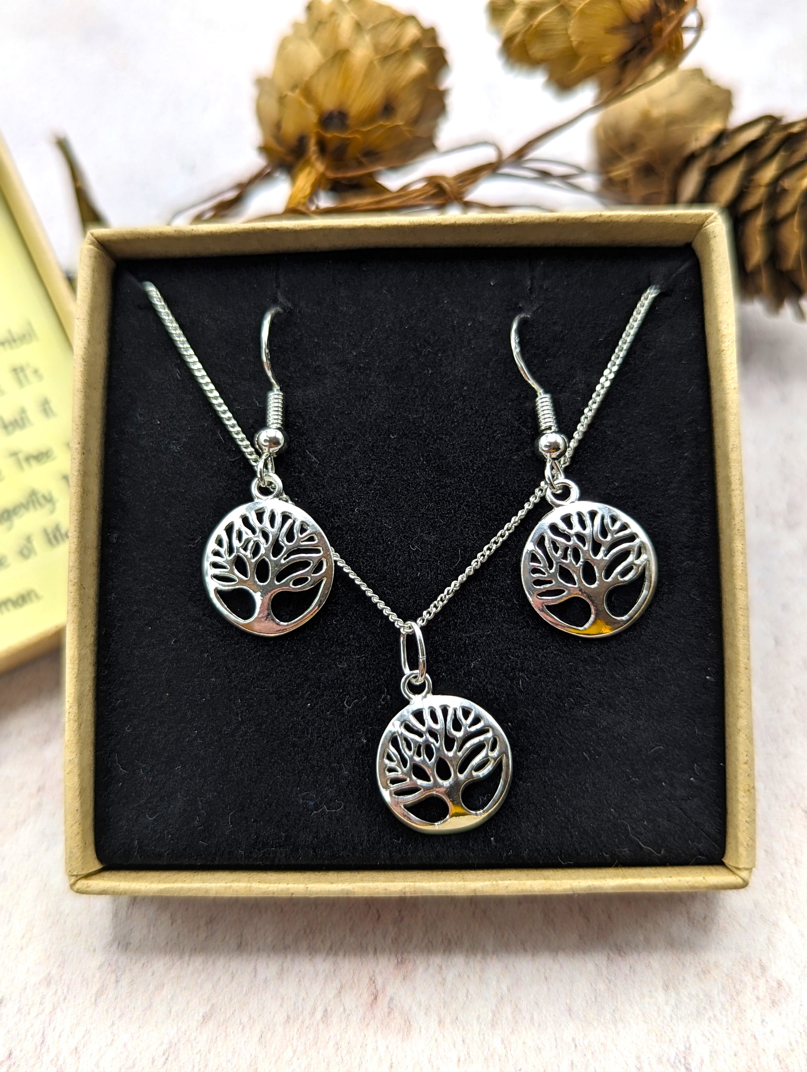The Silver Sunbird Sterling Silver Tree of Life Jewellery Set includes a necklace and matching hypoallergenic earrings, each with a round Tree of Life pendant, beautifully presented in a gift box with dried flowers in the background.