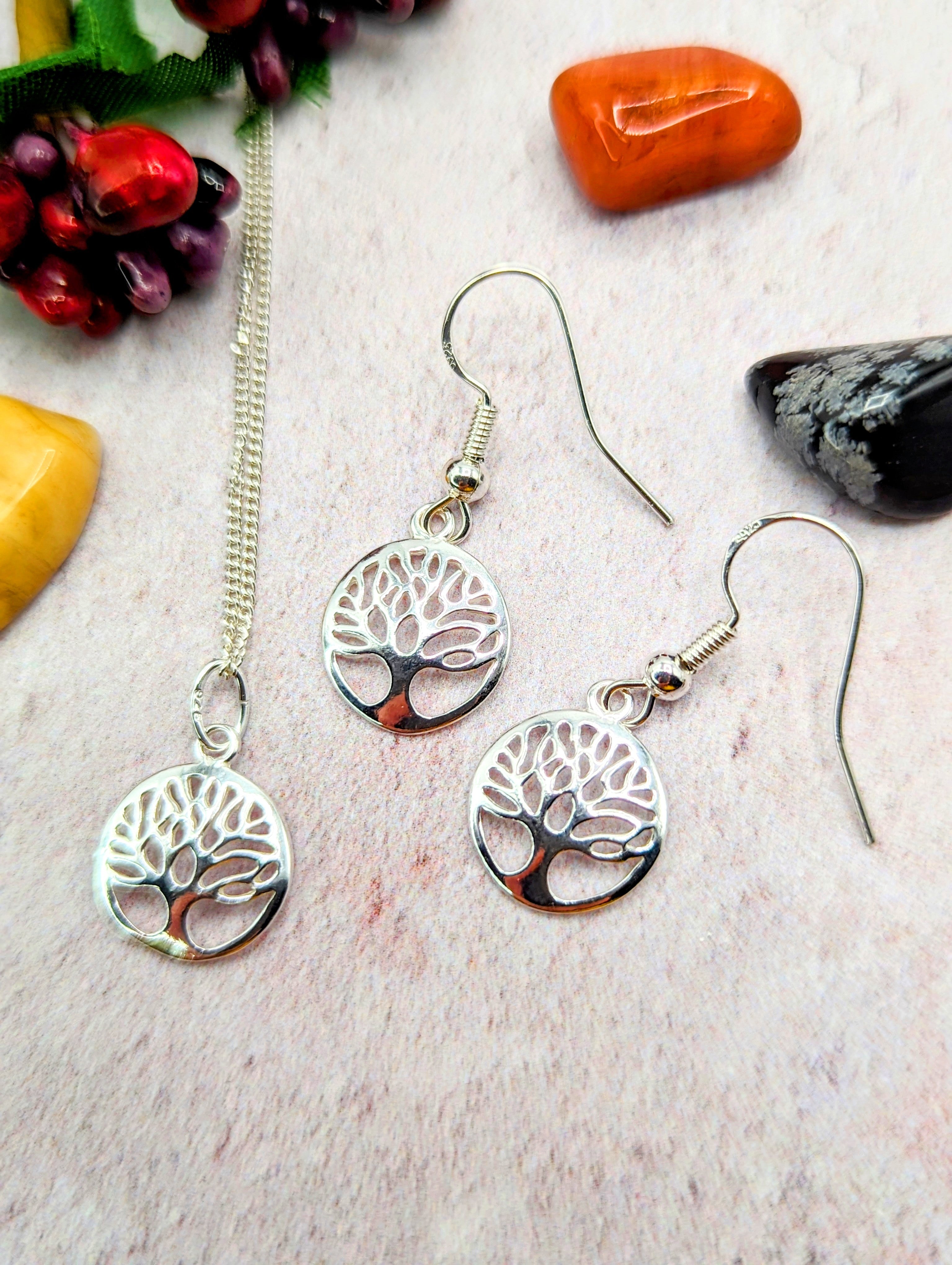 The Silver Sunbird Sterling Silver Tree of Life Jewellery Set – Necklace & Earrings is showcased on a light textured surface with colorful polished stones and decorative berries.
