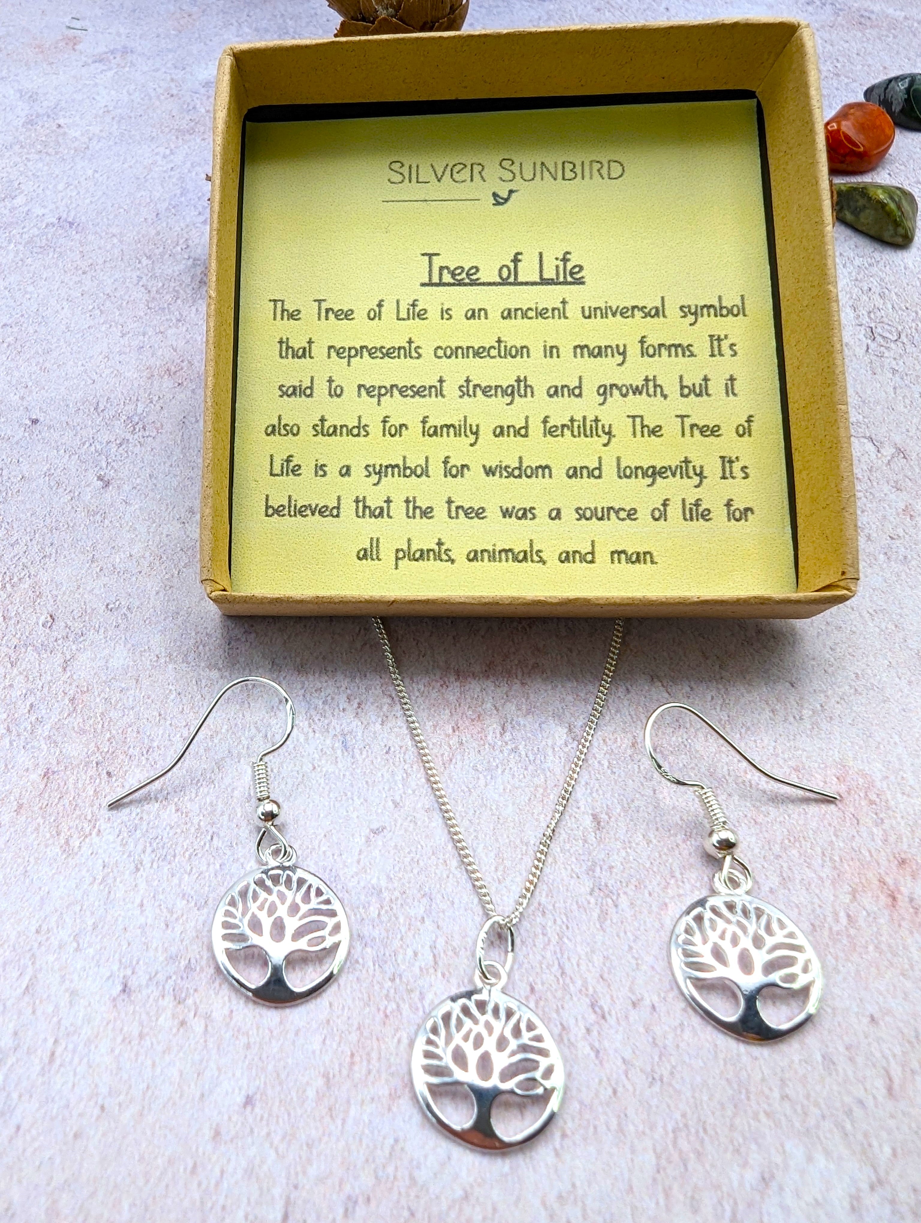 The Sterling Silver Tree of Life Jewellery Set – Necklace & Earrings by Silver Sunbird, featuring hypoallergenic earrings, is displayed on a light surface before a box with a note describing the Tree of Life as a symbol of strength, growth, and wisdom.