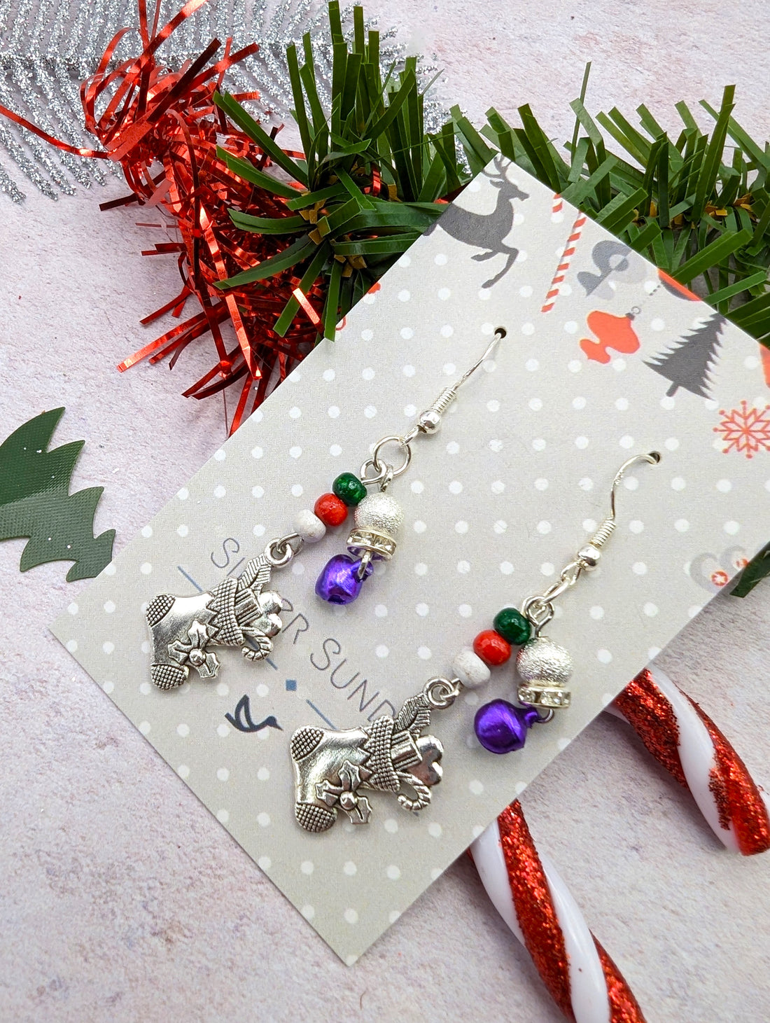 Introducing the Jingle Stockings Earrings by Silver Sunbird, a striking piece of handmade jewelry crafted with sterling silver reindeer and complemented by vibrant red, green, and purple beads. Displayed on a decorative card with candy canes, lush greenery, and red tinsel in the background to create an enchanting holiday-themed setting.