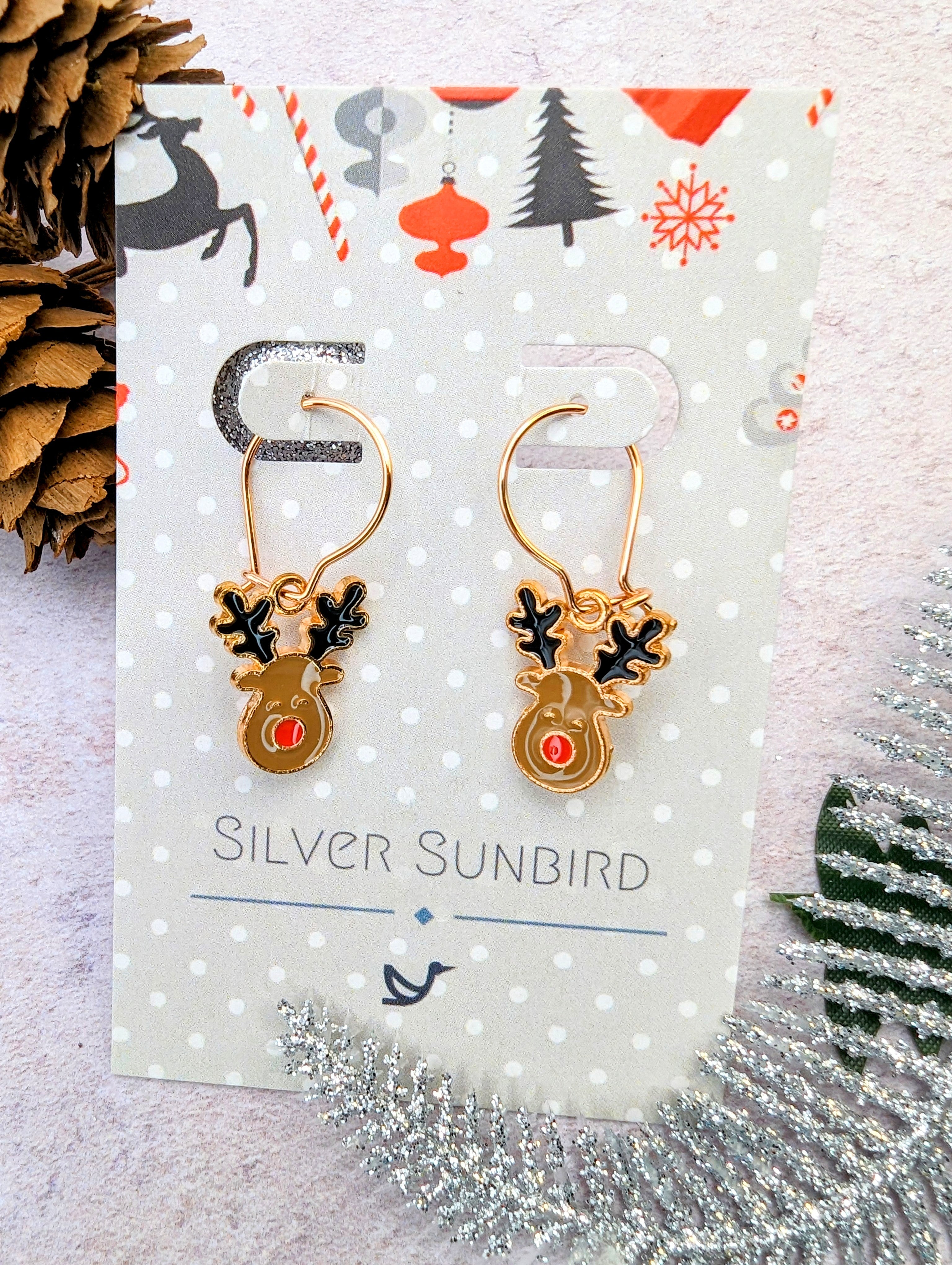 Rosie Reindeer Hoop Earrings with gold finish, red noses, and black antlers are presented on a decorative card from Silver Sunbird. The backdrop includes holiday-themed patterns and pine cones, creating a festive winter ambiance.