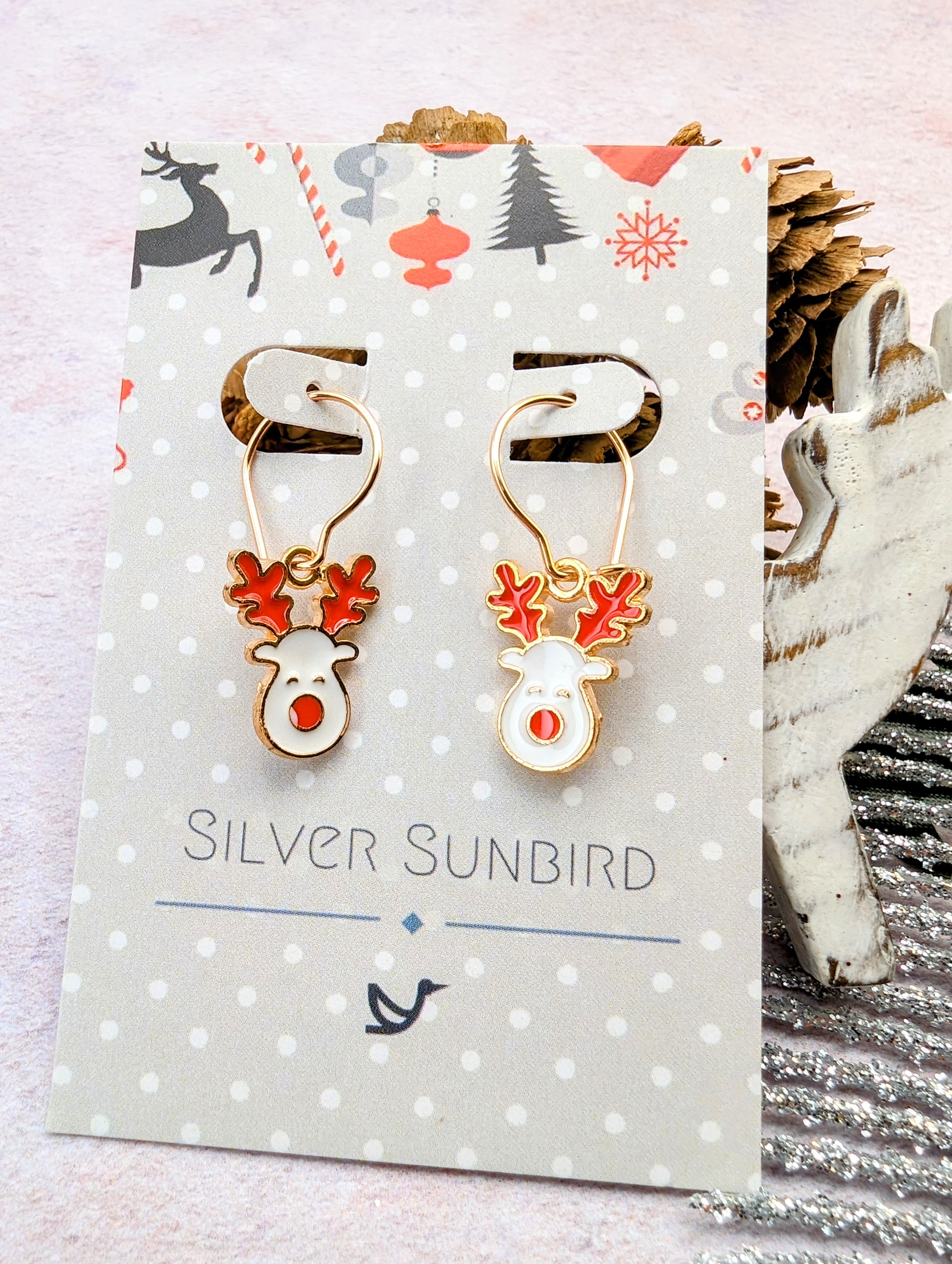 The Rosie Reindeer Hoop Earrings with red antlers and noses are elegantly showcased on a polka dot card branded "Silver Sunbird." Featuring festive enamel designs, the background displays cheerful Christmas motifs and a charming pinecone decoration.