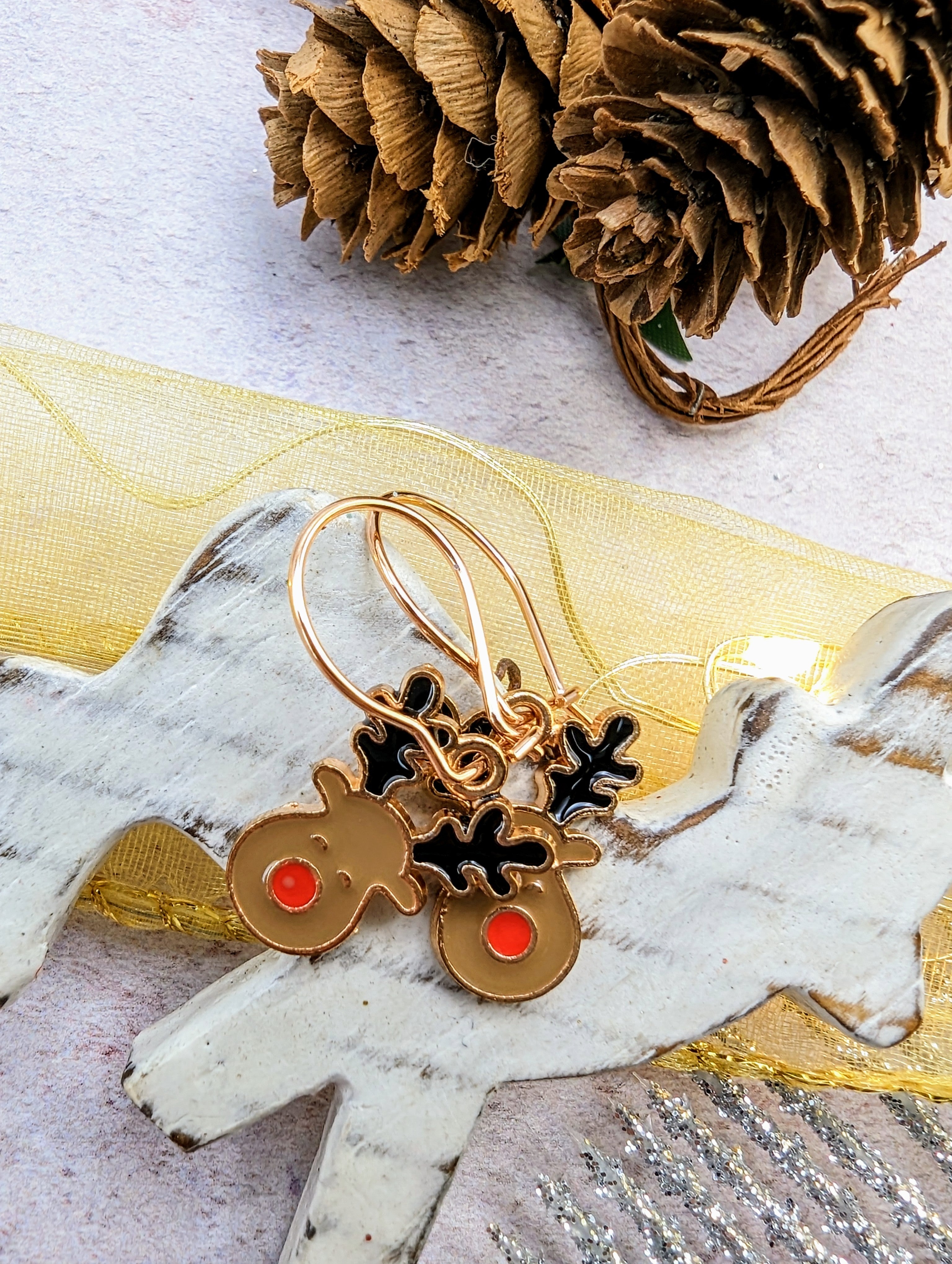 A pair of Rosie Reindeer Hoop Earrings by Silver Sunbird, featuring charming red noses and black antlers, adorn a light wooden reindeer figure. Nearby, a pinecone and a yellow ribbon accentuate the festive atmosphere. These delightful enamel hoop earrings perfectly capture the spirit of the season.