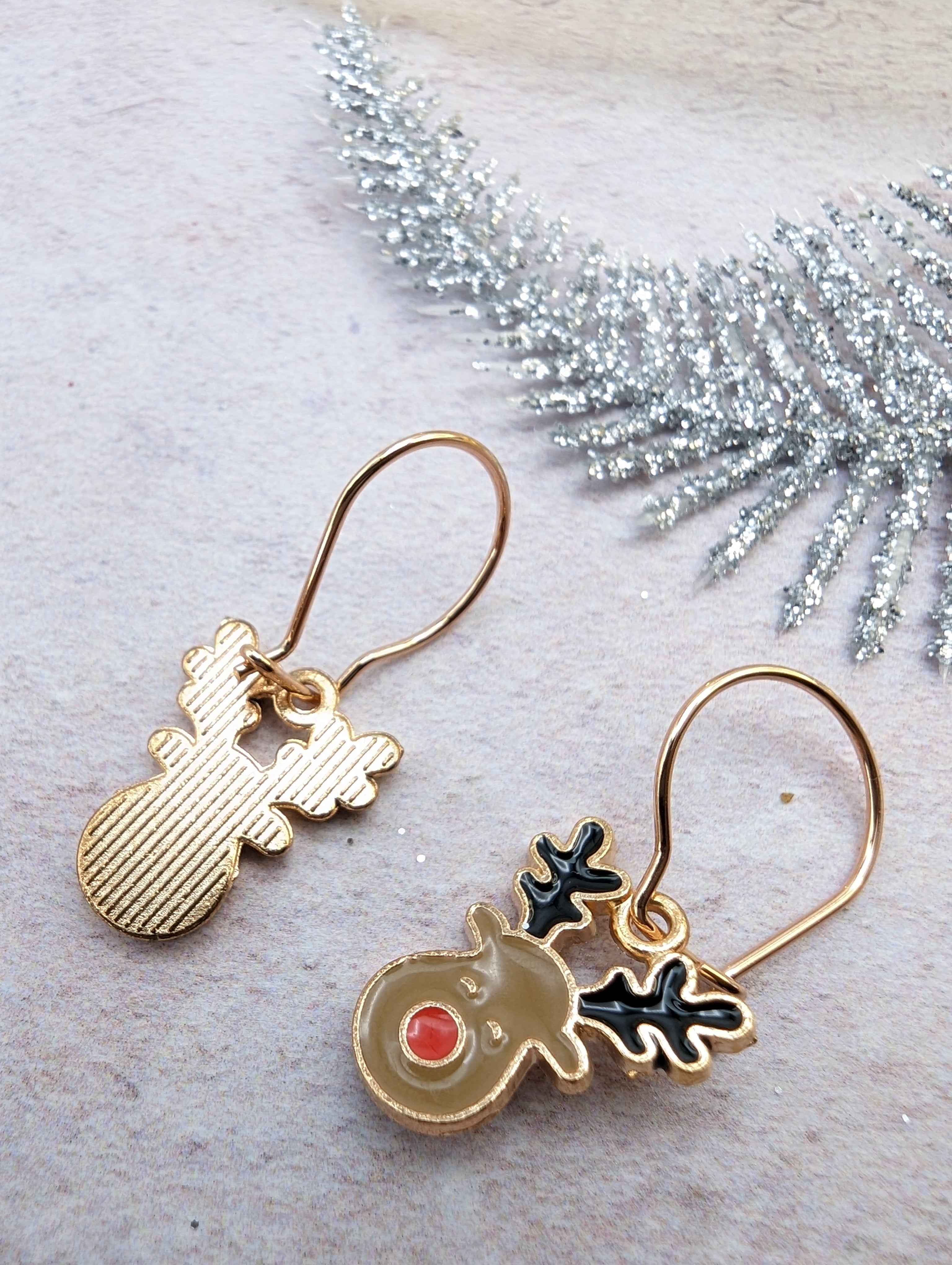 The Rosie Reindeer Hoop Earrings by Silver Sunbird, showcasing festive gold reindeer shapes with black antlers and red noses, are elegantly displayed on a textured surface adorned with a sparkling silver branch.