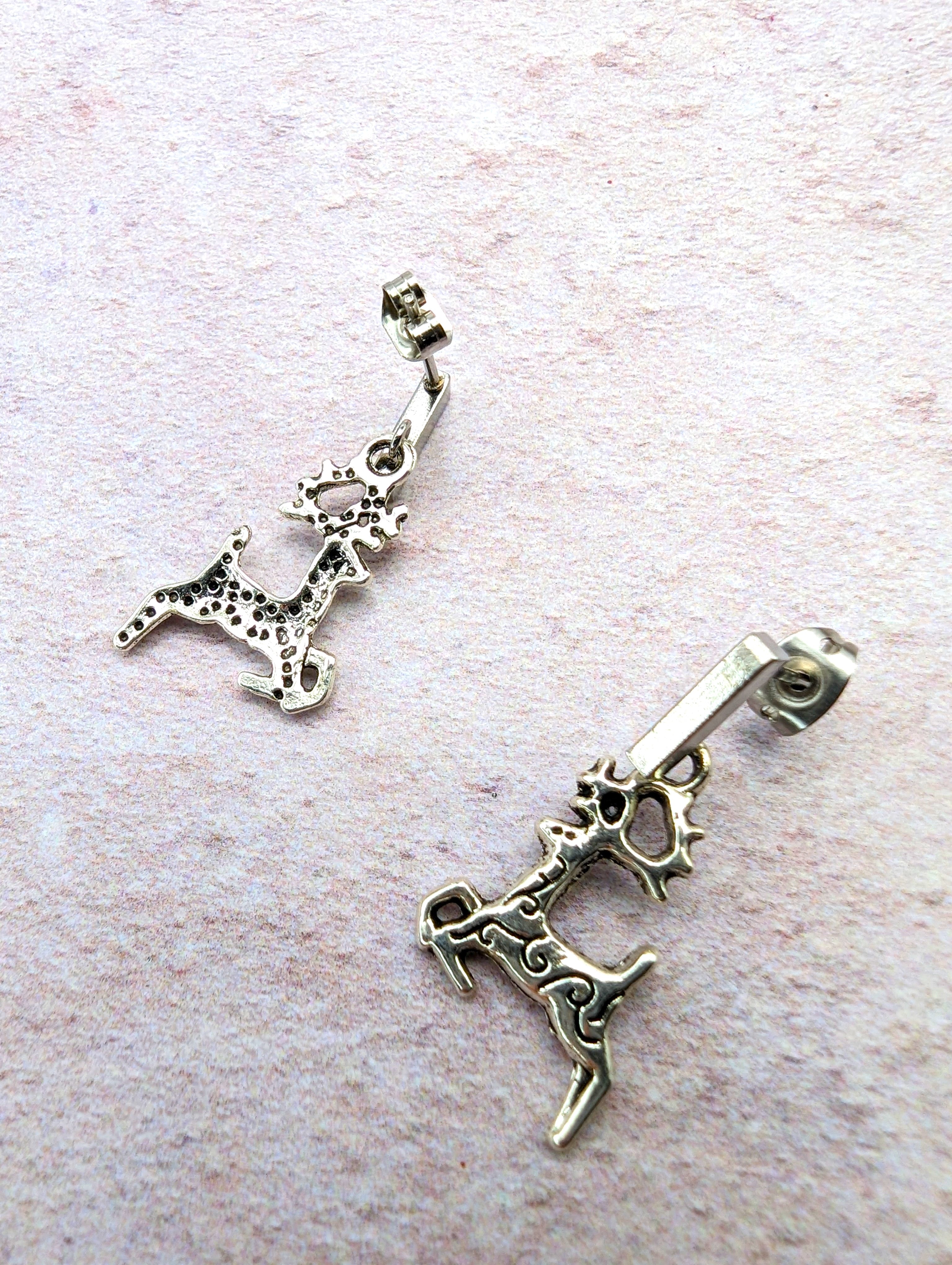 A pair of Prancing Reindeer Earrings from Silver Sunbird, crafted from stainless steel with a rustic, antique finish and intricate detailing, lies on a textured, light-colored surface. These playful and dynamic earrings add charm to your collection of handmade jewelry.