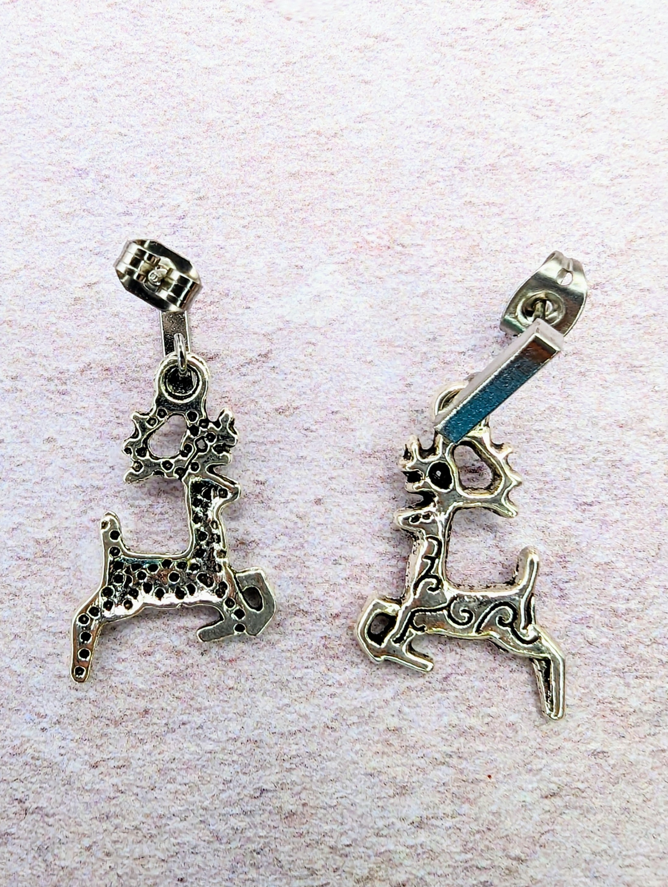 On a textured light pink surface, two Prancing Reindeer Earrings from Silver Sunbird are displayed. Each handmade silver earring showcases unique and intricate patterns, with one featuring a dotted design and the other a swirled pattern. Both pieces include antlers and a bail on top for hanging, highlighting exquisite craftsmanship in jewelry design.