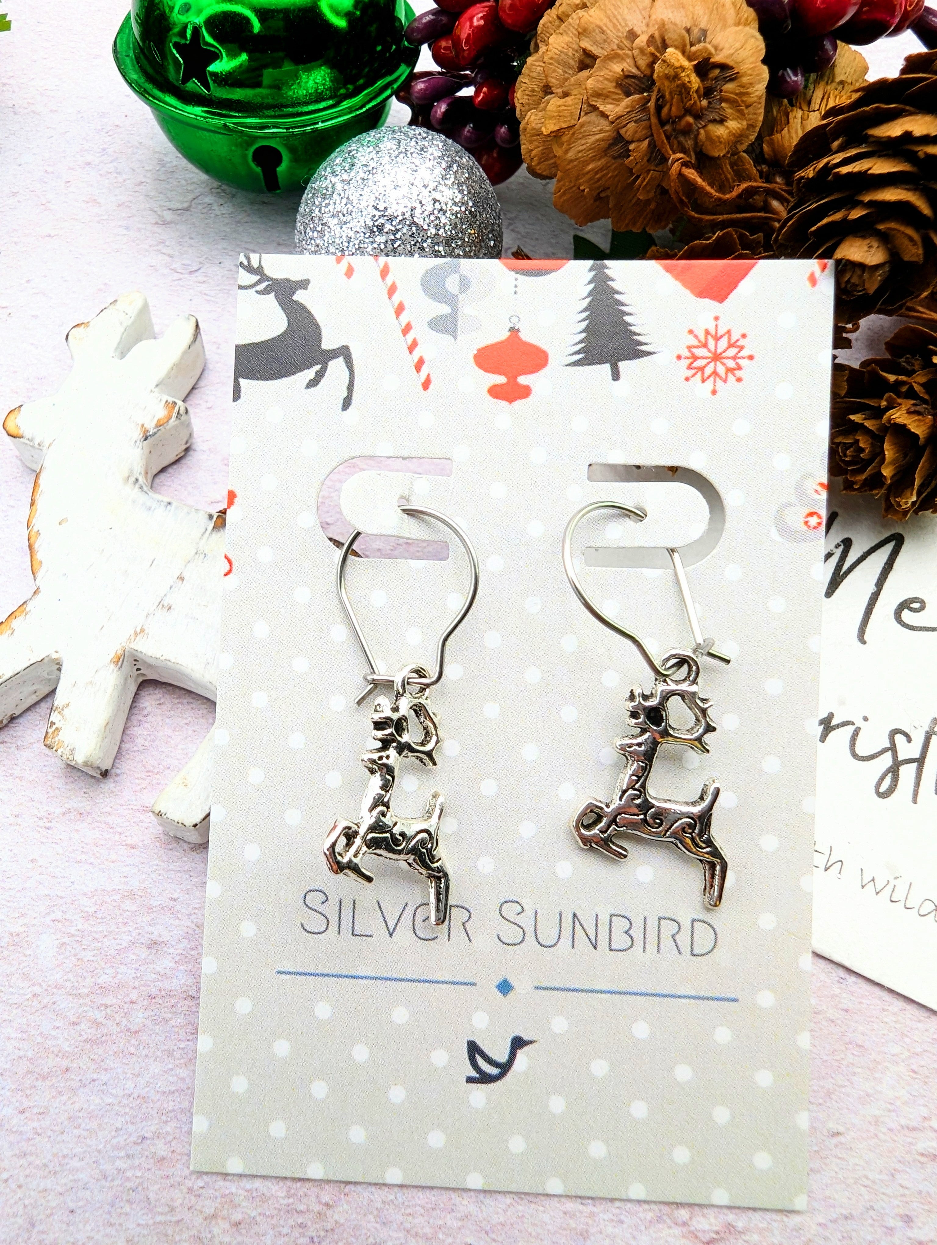 The Prancing Reindeer Earrings by Silver Sunbird are presented on a celebratory card featuring a Christmas motif. The backdrop showcases a green bauble, pinecone, and red berries for a festive theme. Crafted from stainless steel, these earrings add an element of handmade jewelry elegance to any holiday ensemble.