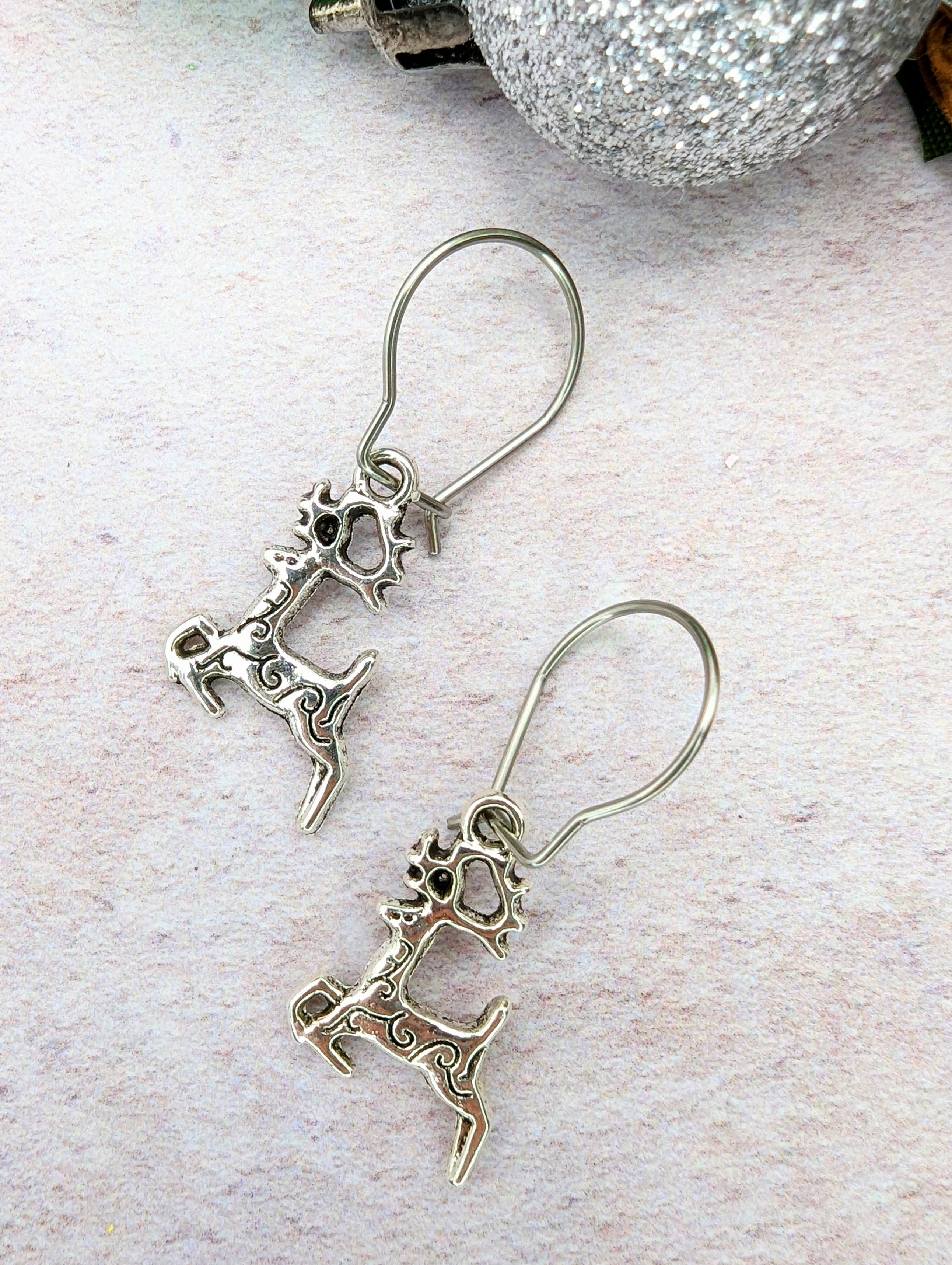 Prancing Reindeer Earrings by Silver Sunbird are displayed on a textured surface alongside a sparkling silver ornament.