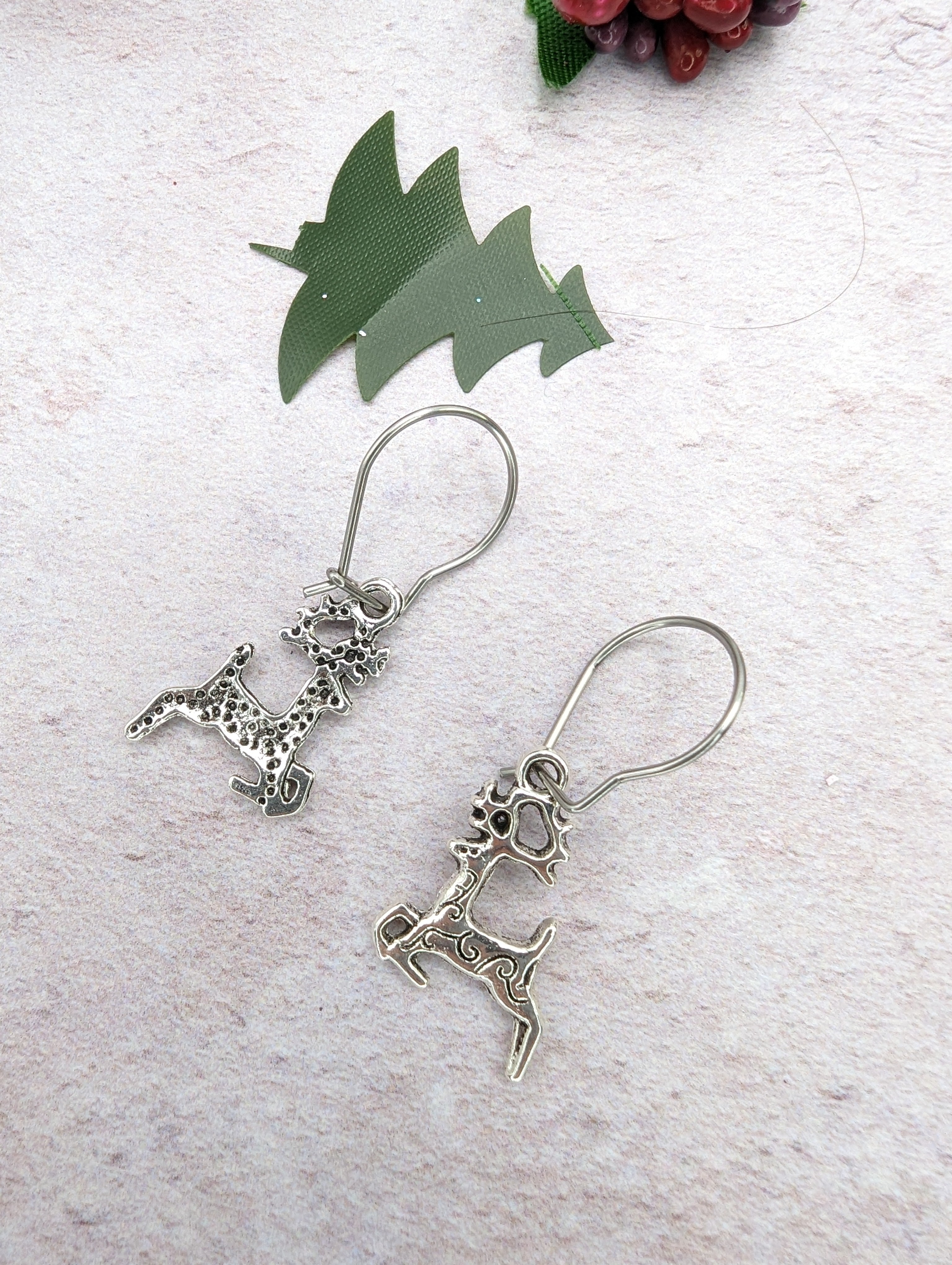 The Prancing Reindeer Earrings by Silver Sunbird, crafted from stainless steel with intricate designs, rest on a light textured surface. Above them, a small green paper cutout shaped like trees and a cluster of small red berries add to their charm.