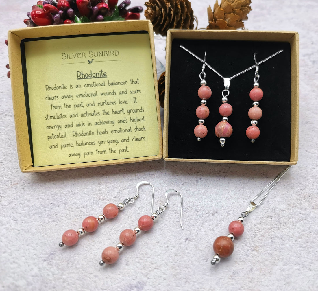 The Silver Sunbird Rhodonite Gemstone Jewellery Set graces the table with a sterling silver necklace, earrings, and bracelet. A card explaining rhodonite&