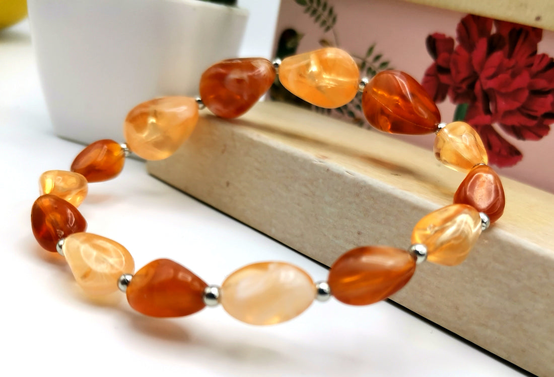 The Silver Sunbird Amber Resin Bracelet, featuring polished, irregularly shaped reclaimed amber resin stones paired with silver spacer beads, is elegantly presented on a wooden surface. A potted plant and a floral-patterned box in the background enhance the charm of the scene.
