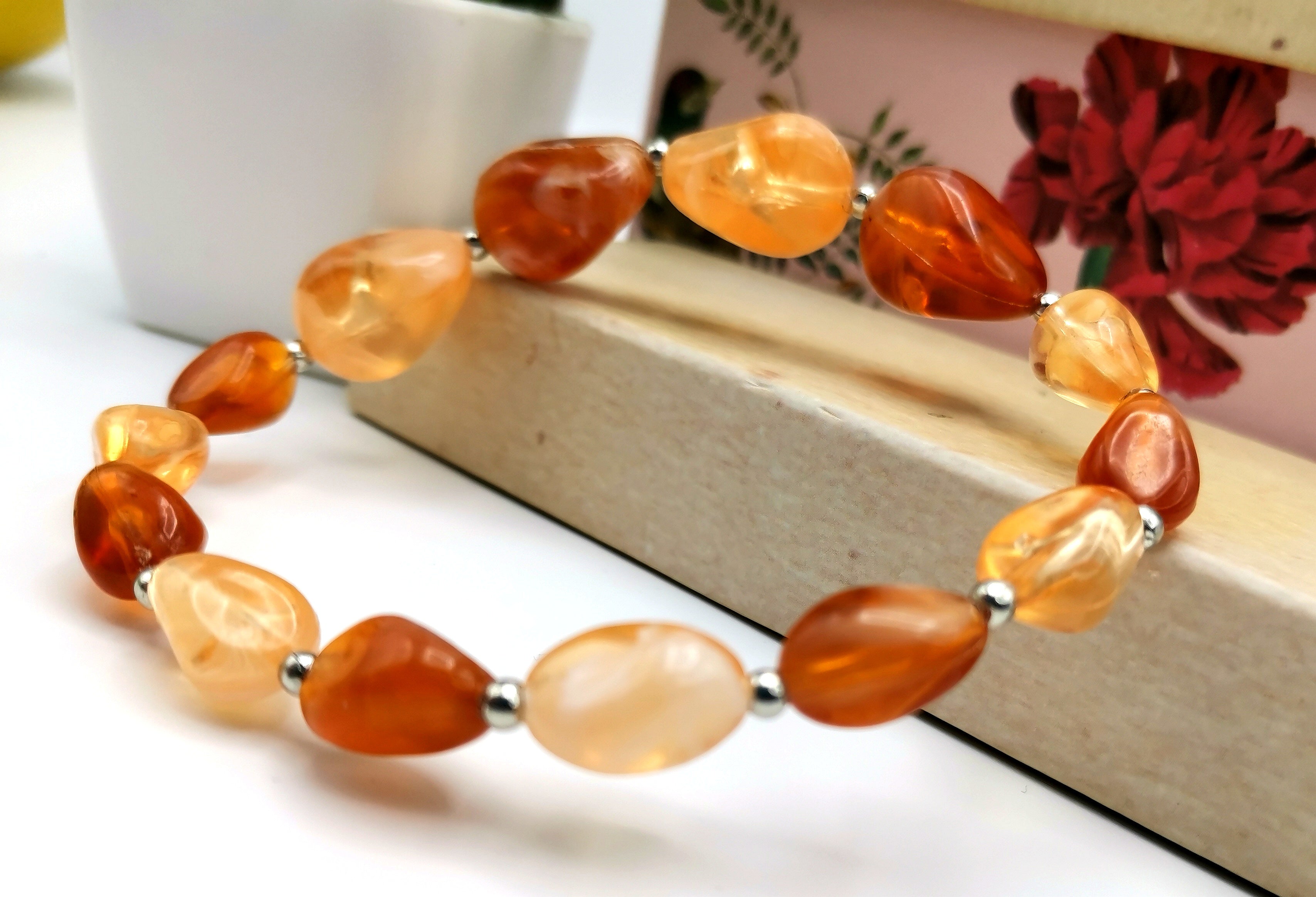 The Silver Sunbird Amber Resin Bracelet, featuring polished, irregularly shaped reclaimed amber resin stones paired with silver spacer beads, is elegantly presented on a wooden surface. A potted plant and a floral-patterned box in the background enhance the charm of the scene.