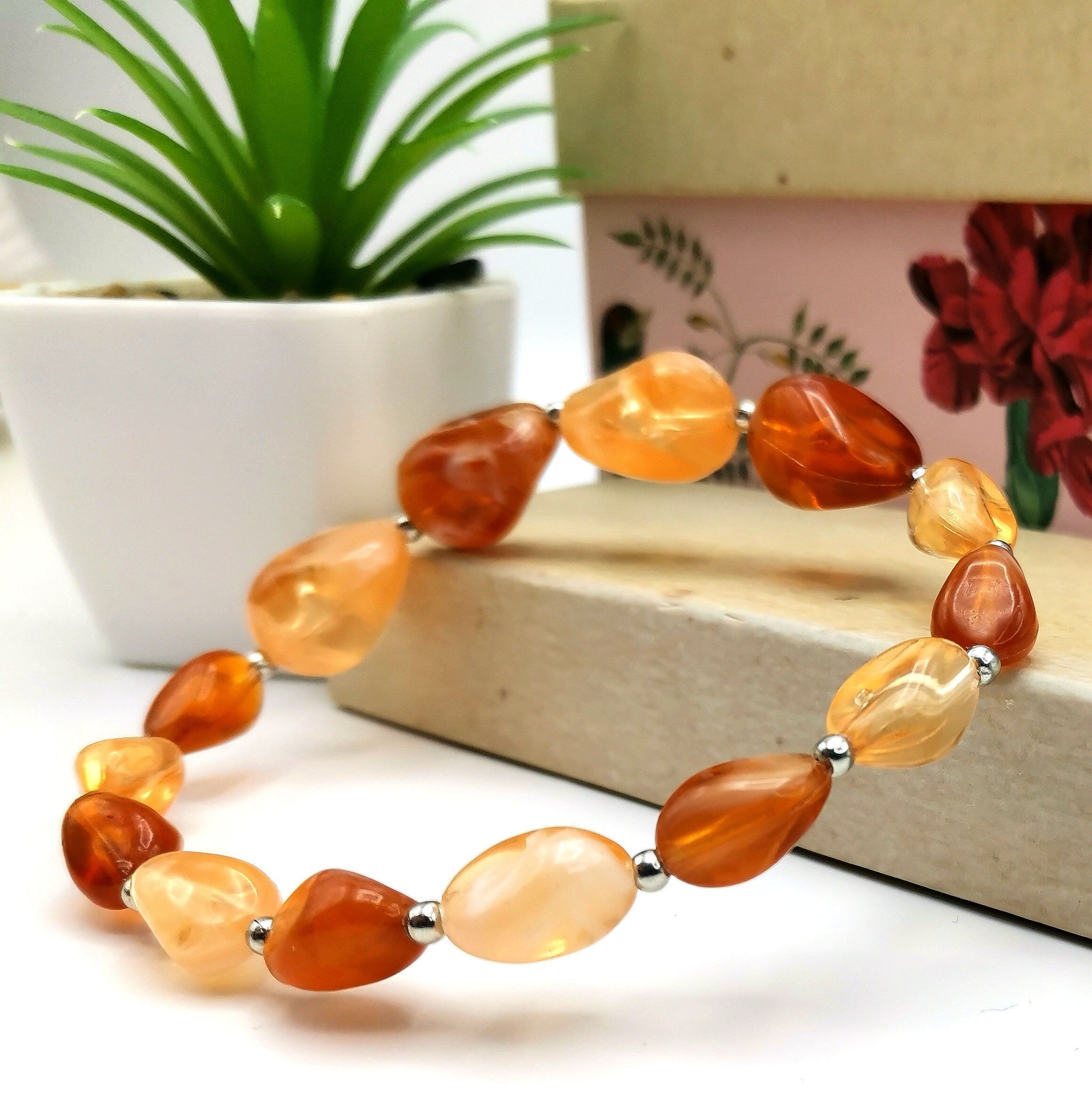 The "Amber Resin Bracelet" by Silver Sunbird, featuring polished orange and peach beads complemented by silver spacer beads, is elegantly displayed on a wooden surface beside a potted green plant.
